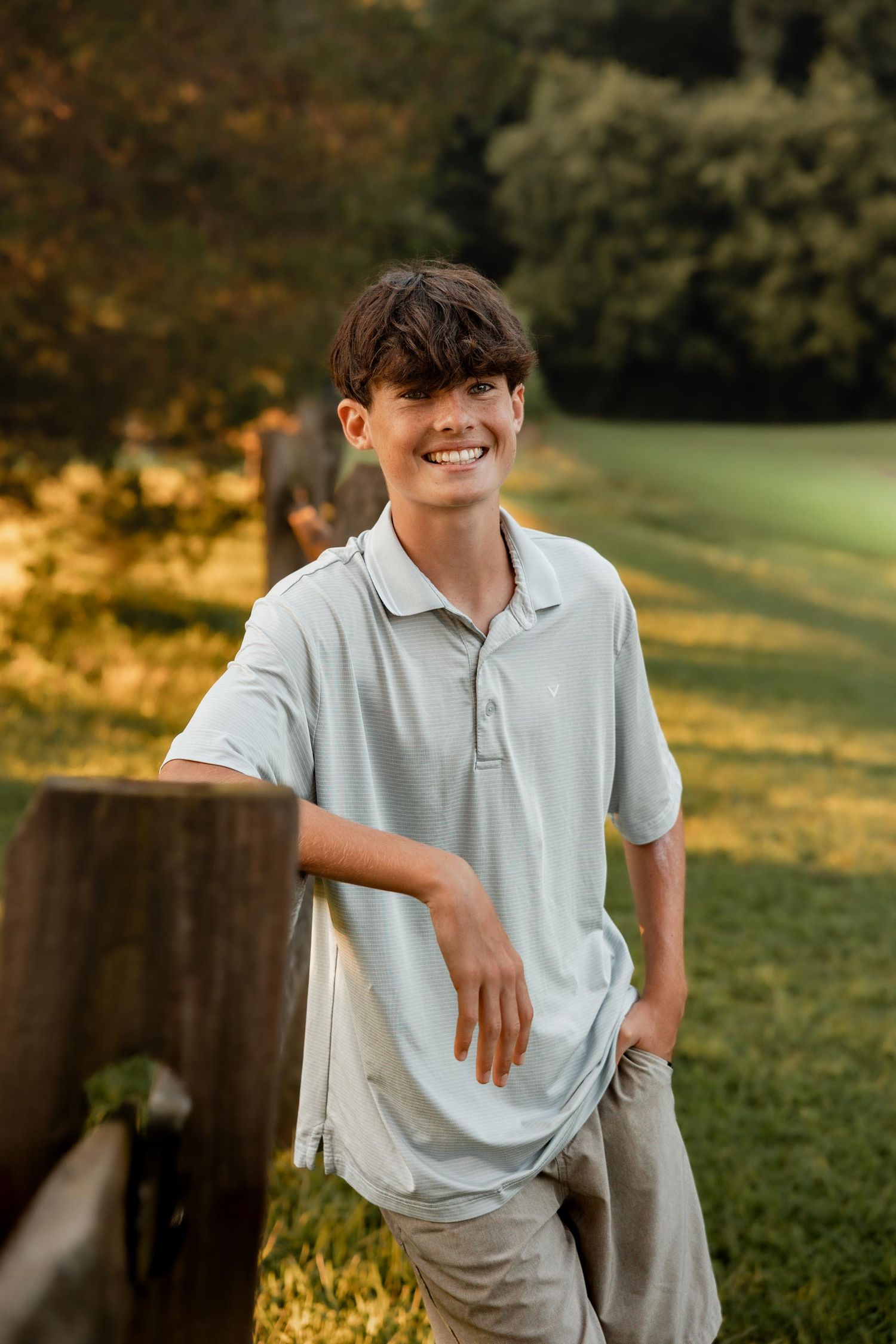 Smiling teenage boy in light blue polo shirt leaning on wooden fence post in sunny green park setting.