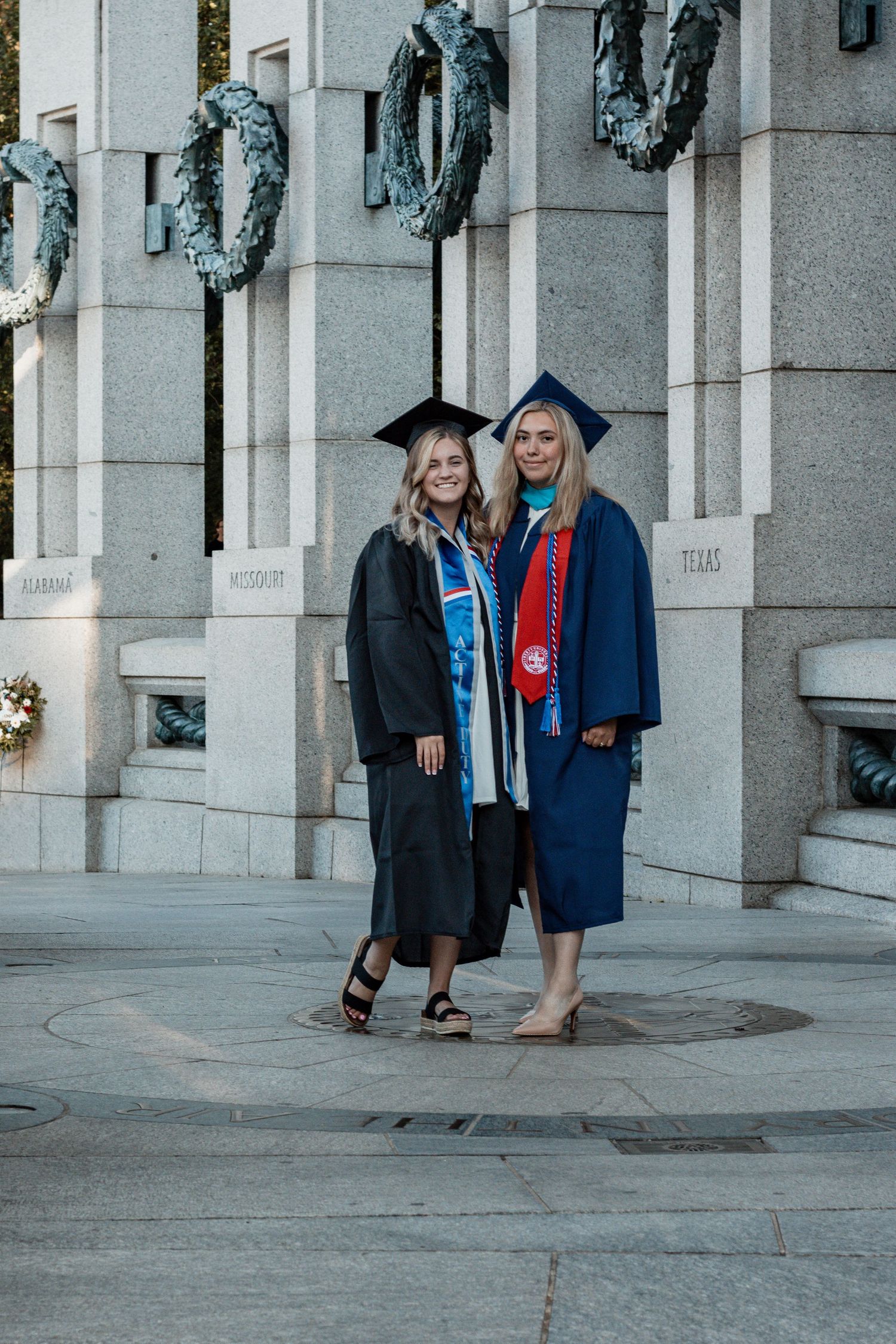 Recent graduates in blue caps and gowns gather around stone pillars on a sunny graduation day.