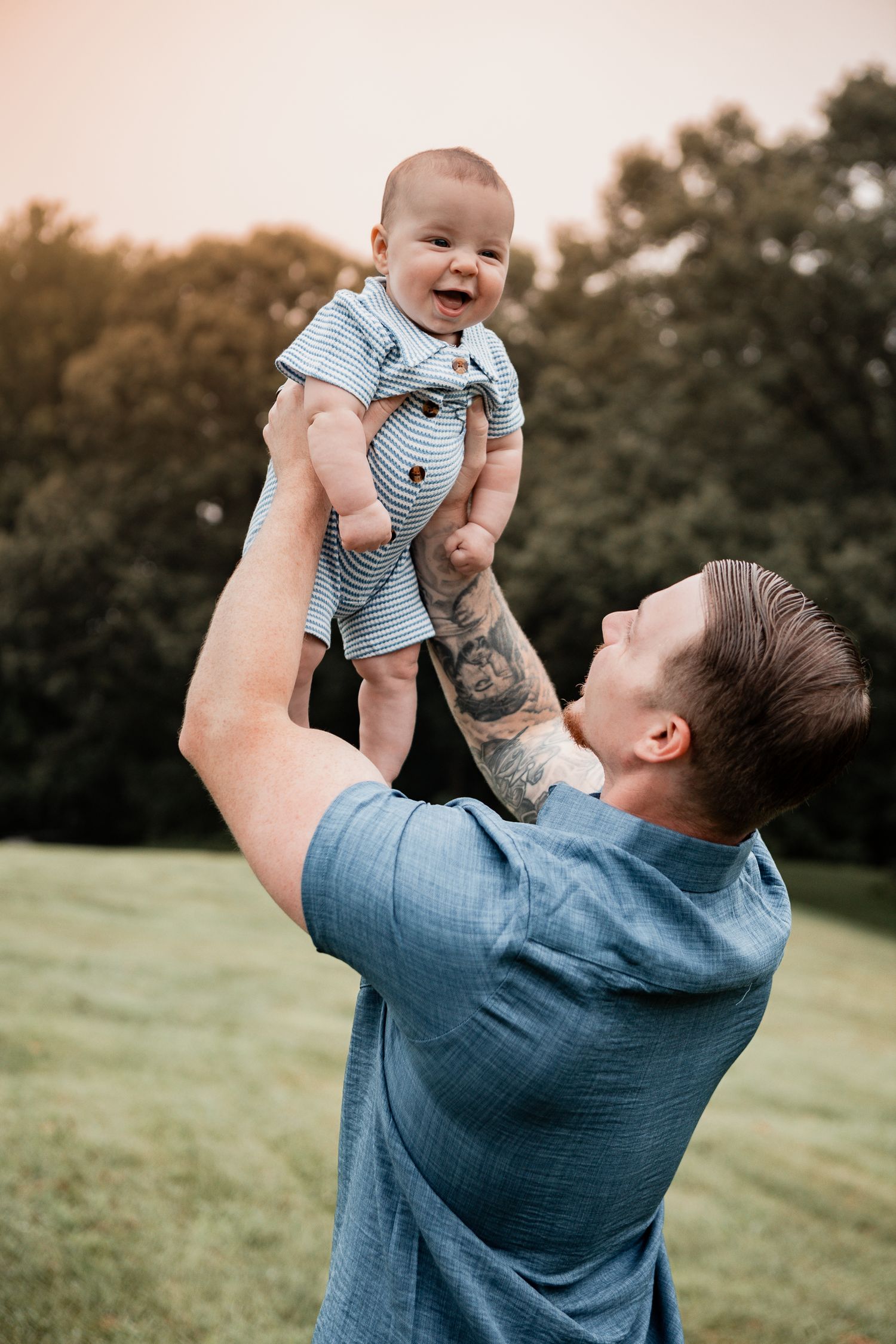 A parent lifts their baby in a blue onesie above their head in a grassy field at sunset creating a joyful moment.