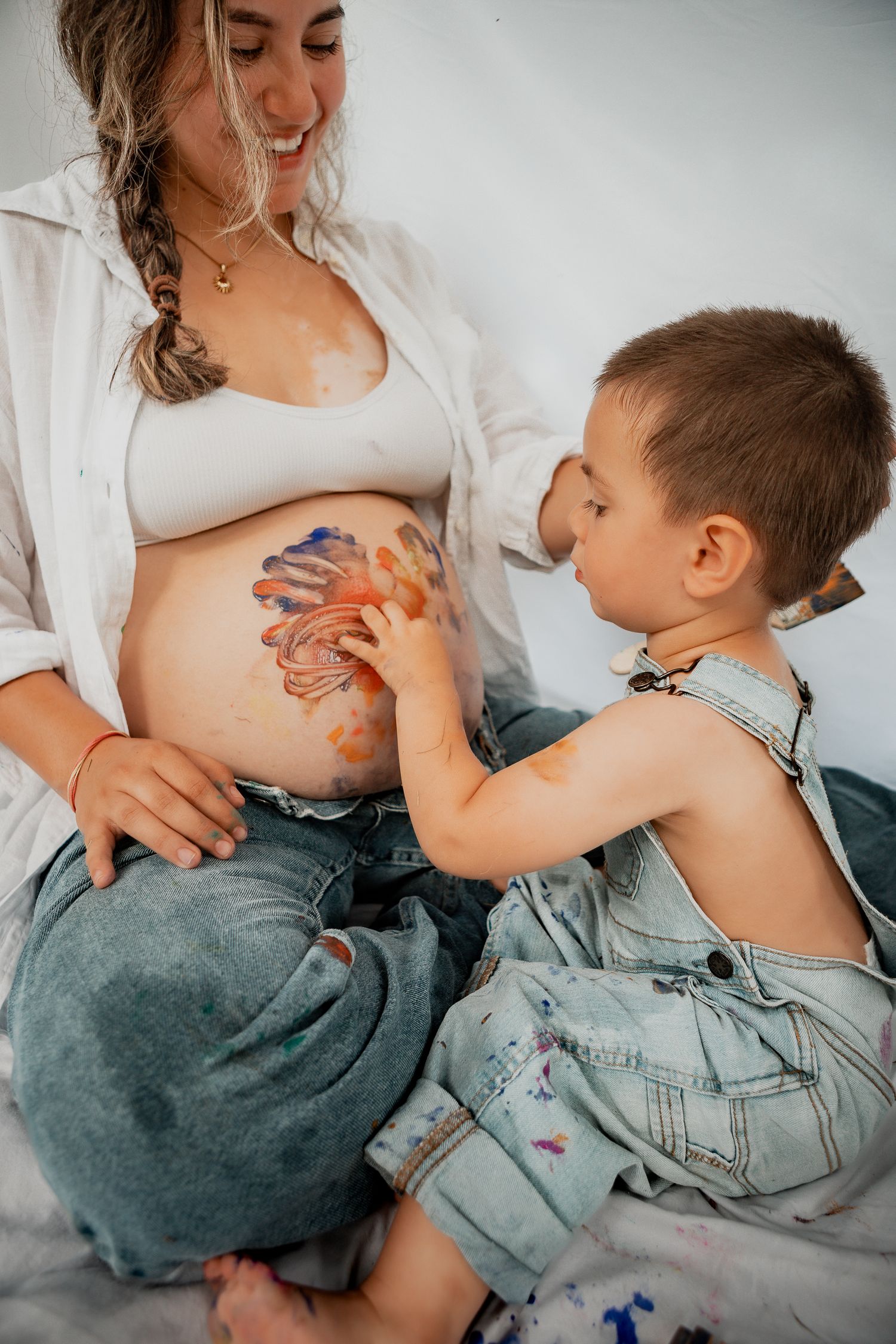 A pregnant woman in a white cardigan smiles while a young child places painted hands on her belly.