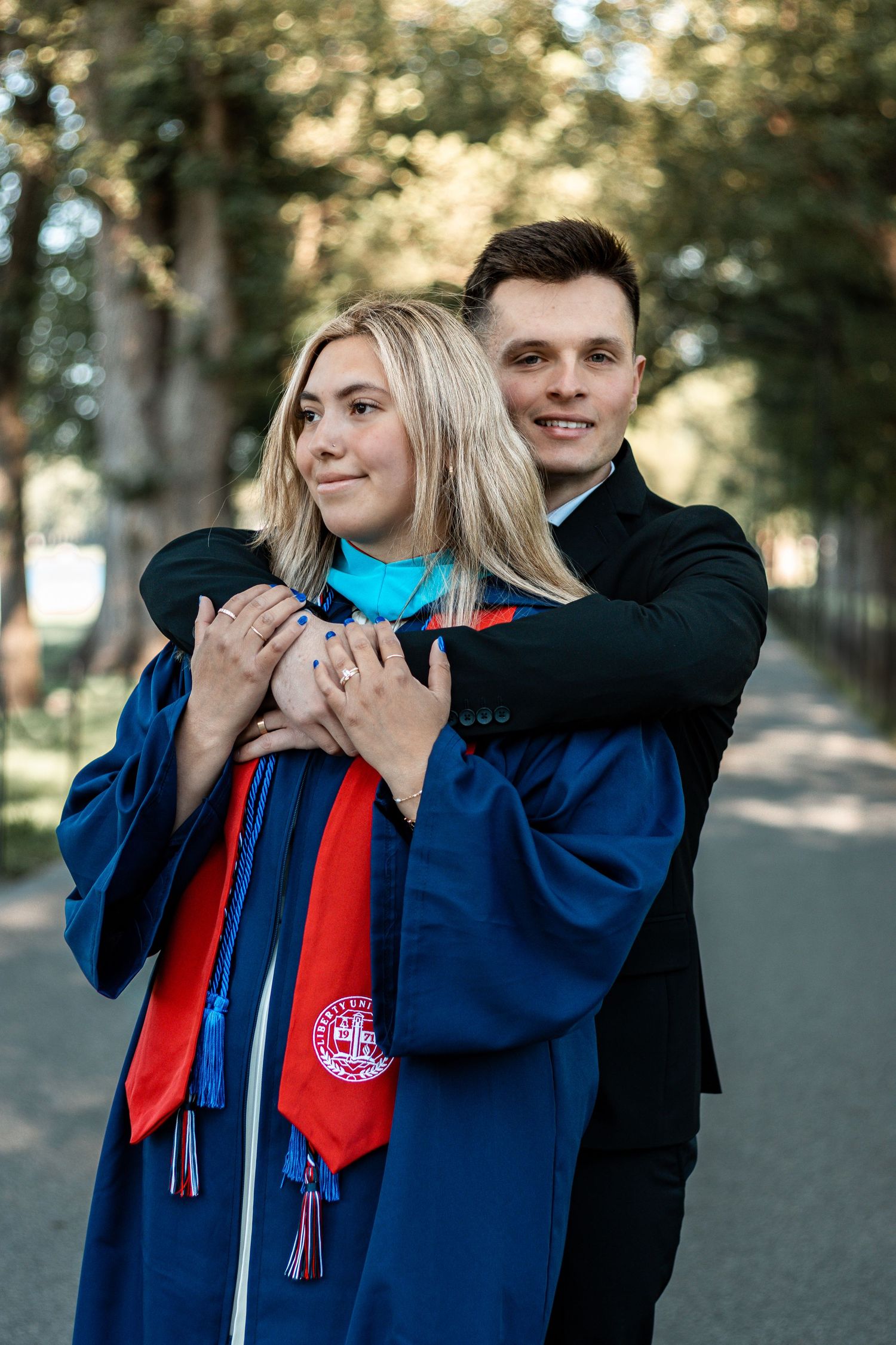 Couple wearing graduation regalia embracing outdoors on tree-lined path.