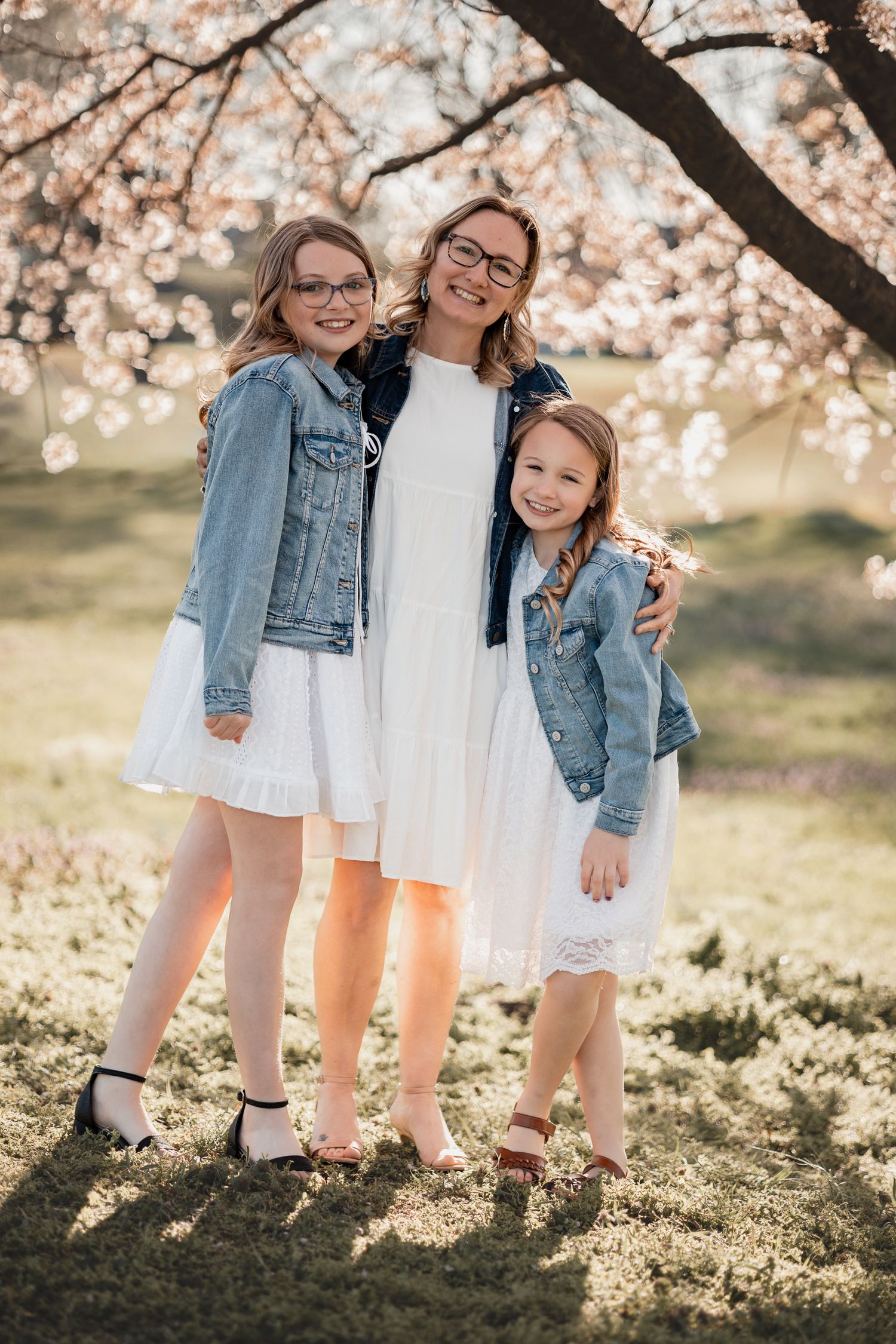 A mother and two daughters in white dresses and denim jackets pose under blooming cherry blossom trees in spring.