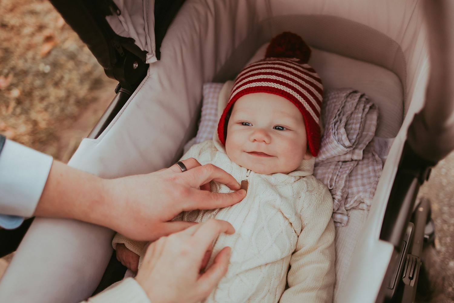 Baby wearing red and white striped knit hat lies in stroller while being gently touched by adult hand.
