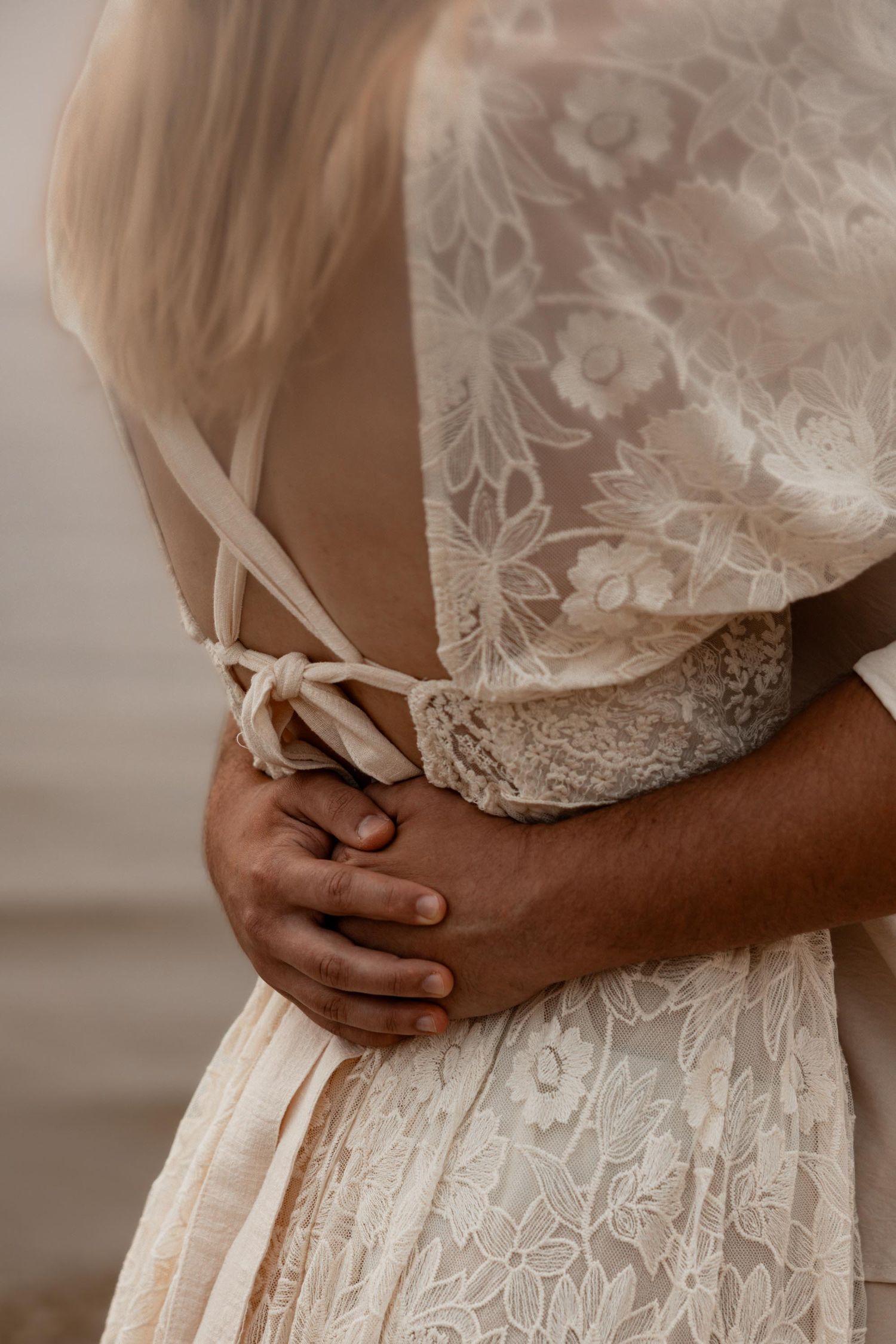 Close-up of a couple embracing, showcasing a beautiful floral lace wedding dress with delicate ribbon tie back detail.