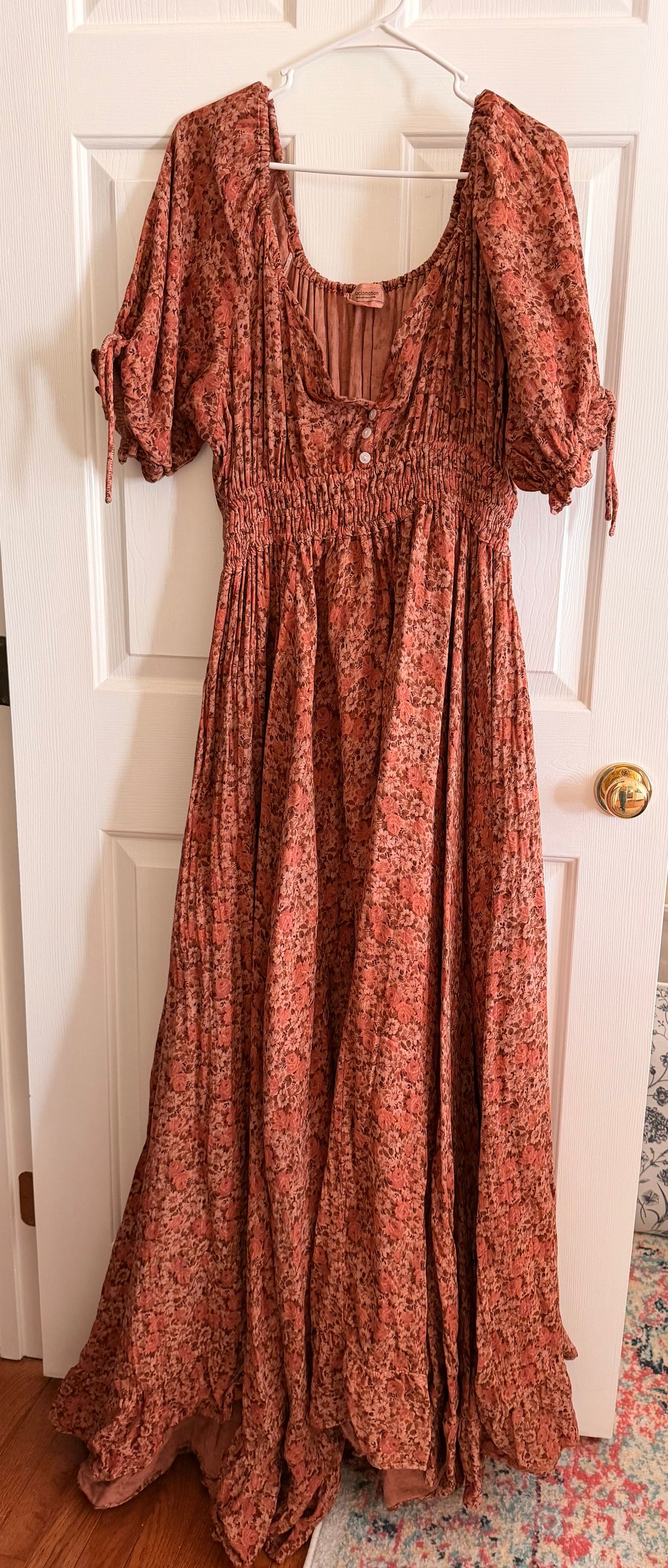 Rust floral maxi dress with smocked bodice, puff sleeves, and tie details, hanging on a white door.