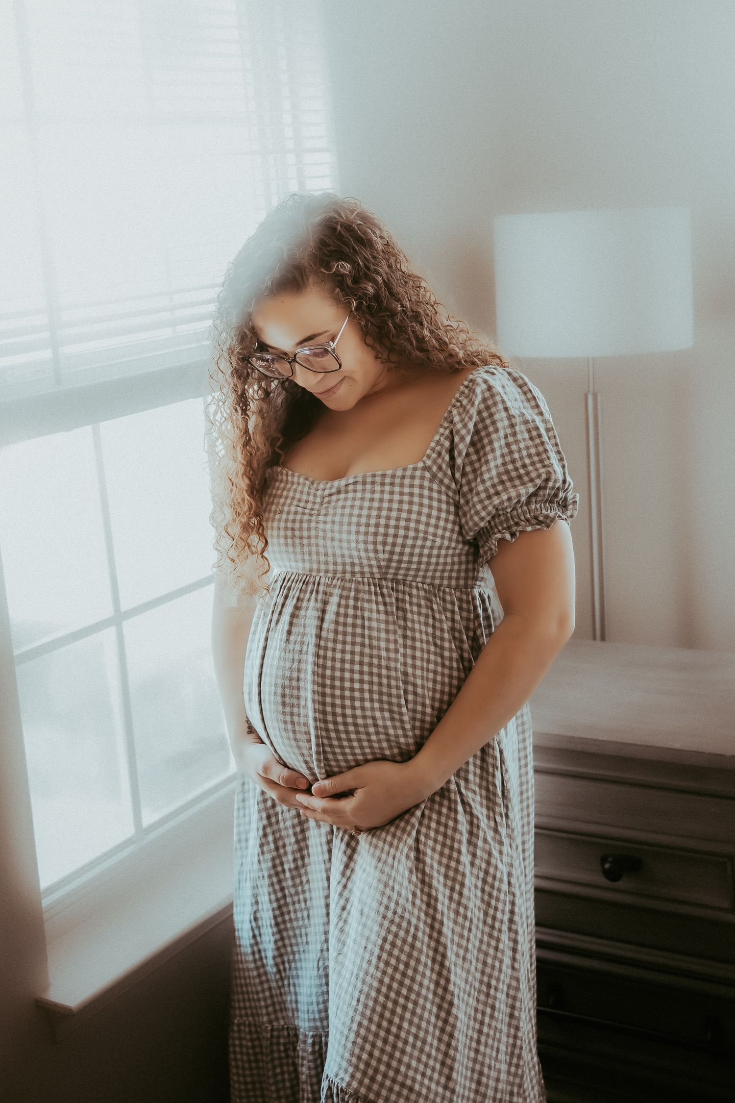 Pregnant woman in gingham dress cradling baby bump near window with soft natural light.
