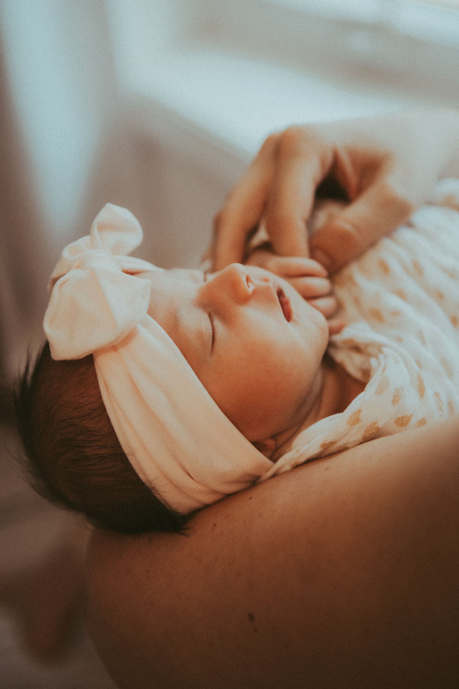 Newborn baby sleeping peacefully in soft white swaddle blanket in warm lighting.