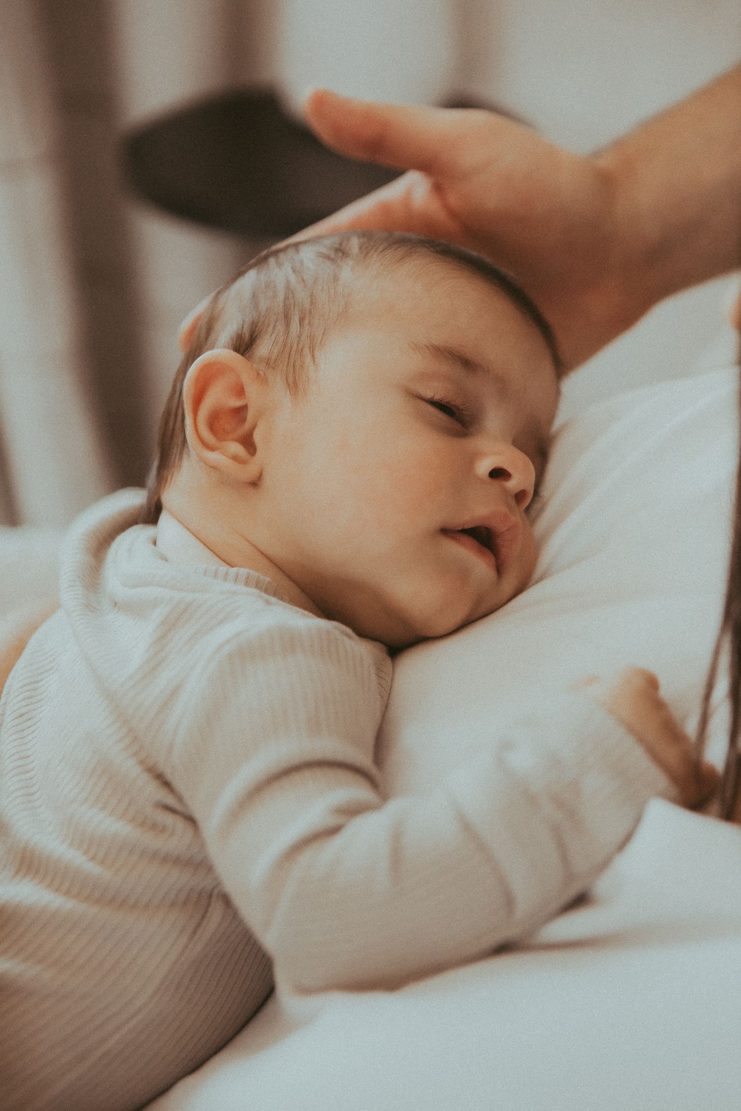 Sleeping baby in white knit clothing rests peacefully against soft bedding in natural light.