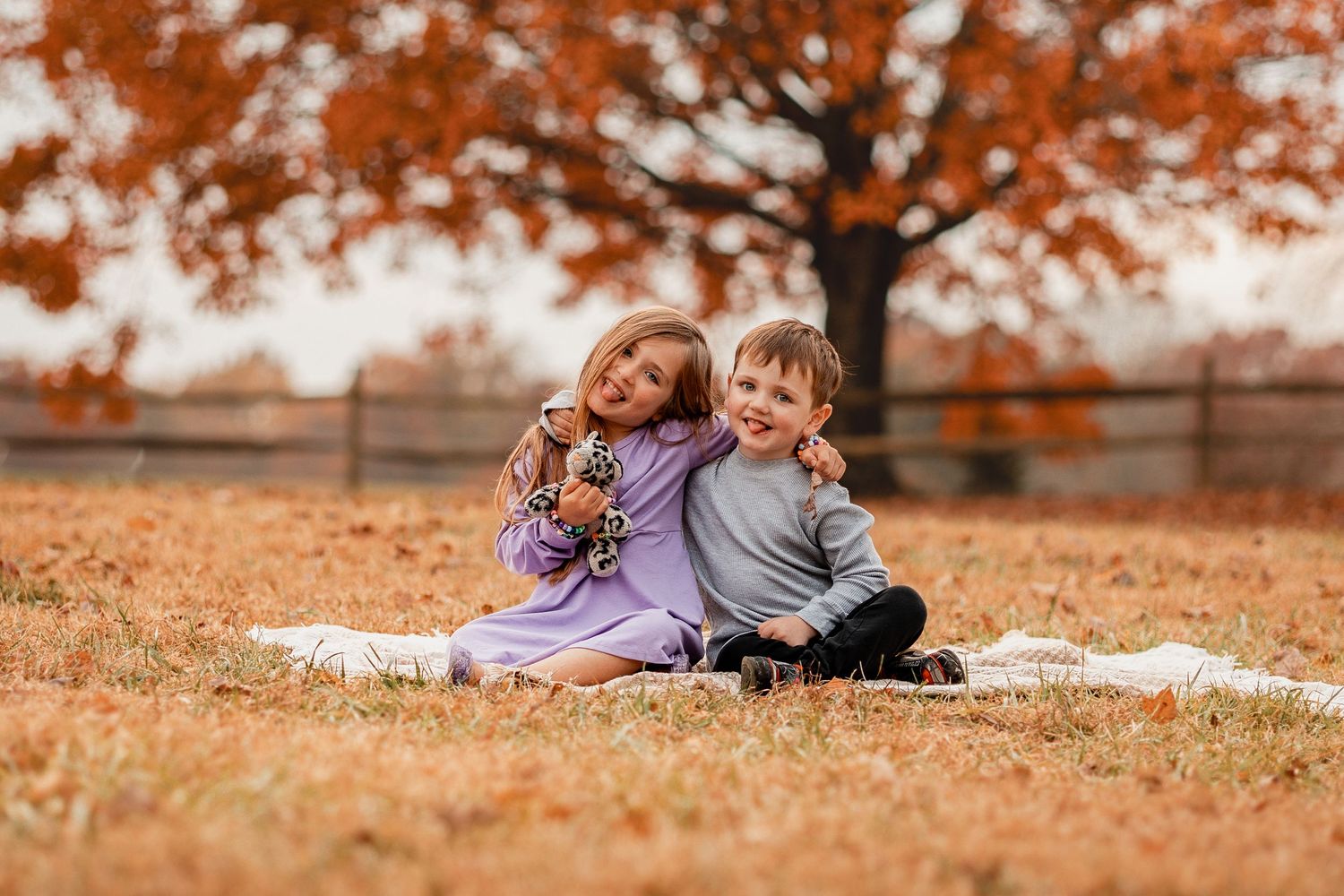 A family relaxes together on a purple blanket surrounded by fallen autumn leaves in a park setting.