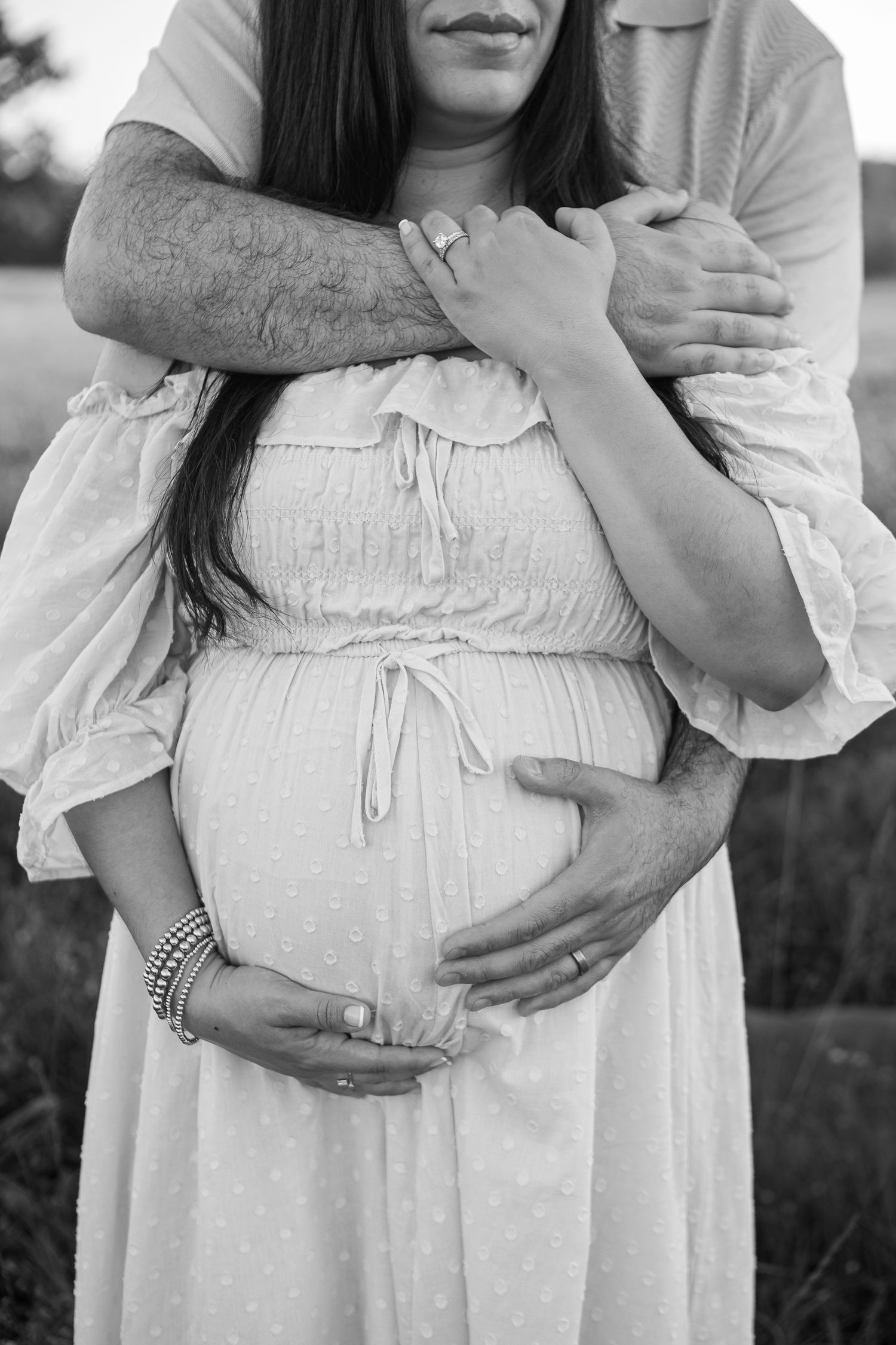 Black and white photo of a couple embracing, showing hands holding a pregnant belly in a flowing white dress.