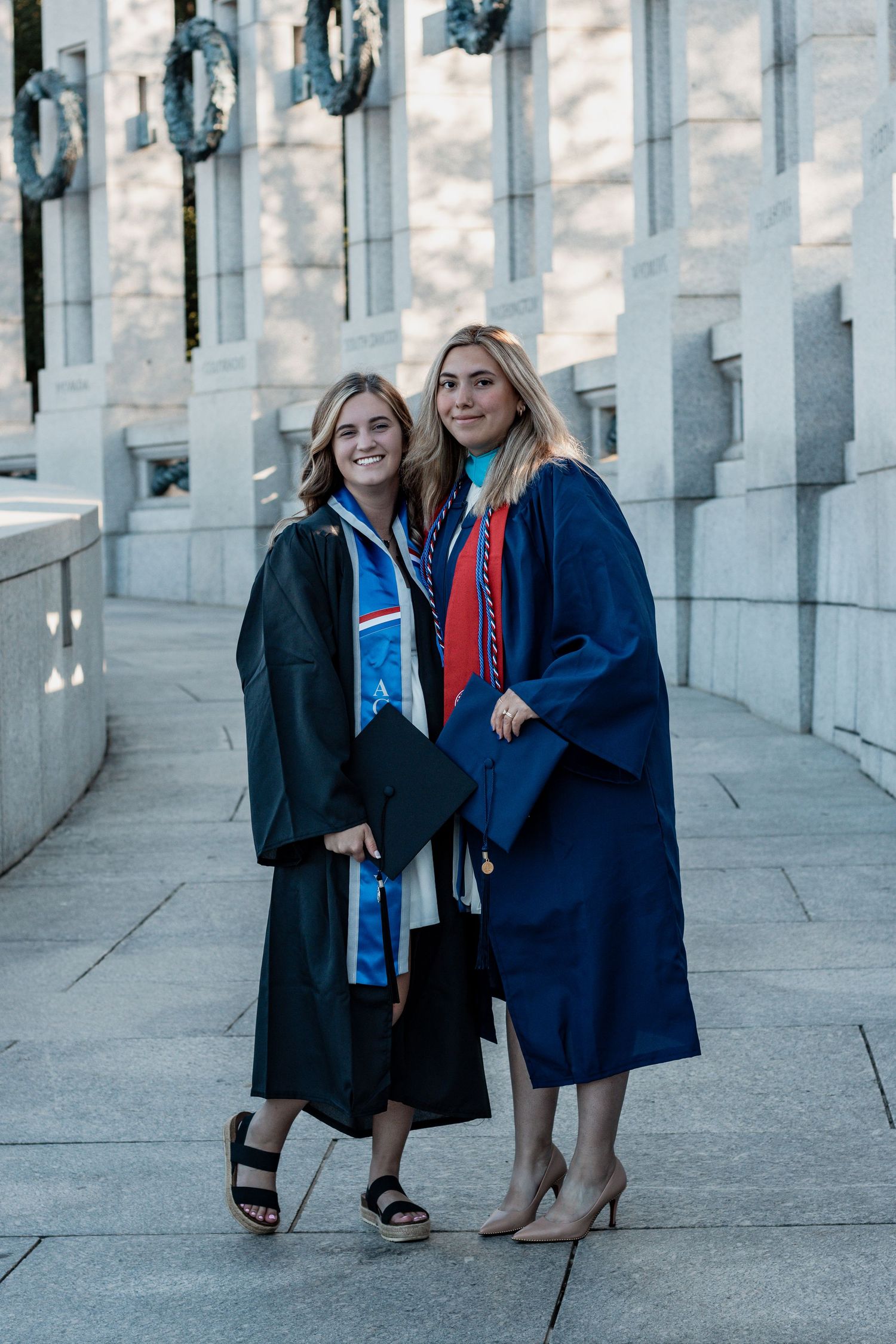Two graduates in academic regalia standing together and smiling in a classical columned architectural setting.