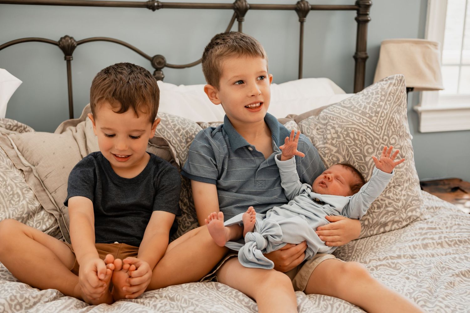 Two children sit on a bed with a sleeping infant propped on pillows in a serene bedroom setting.