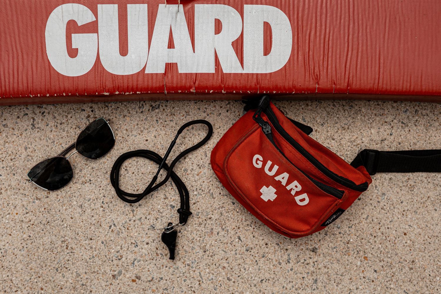 Red lifeguard rescue tubes and sunglasses arranged on concrete pool deck surface.