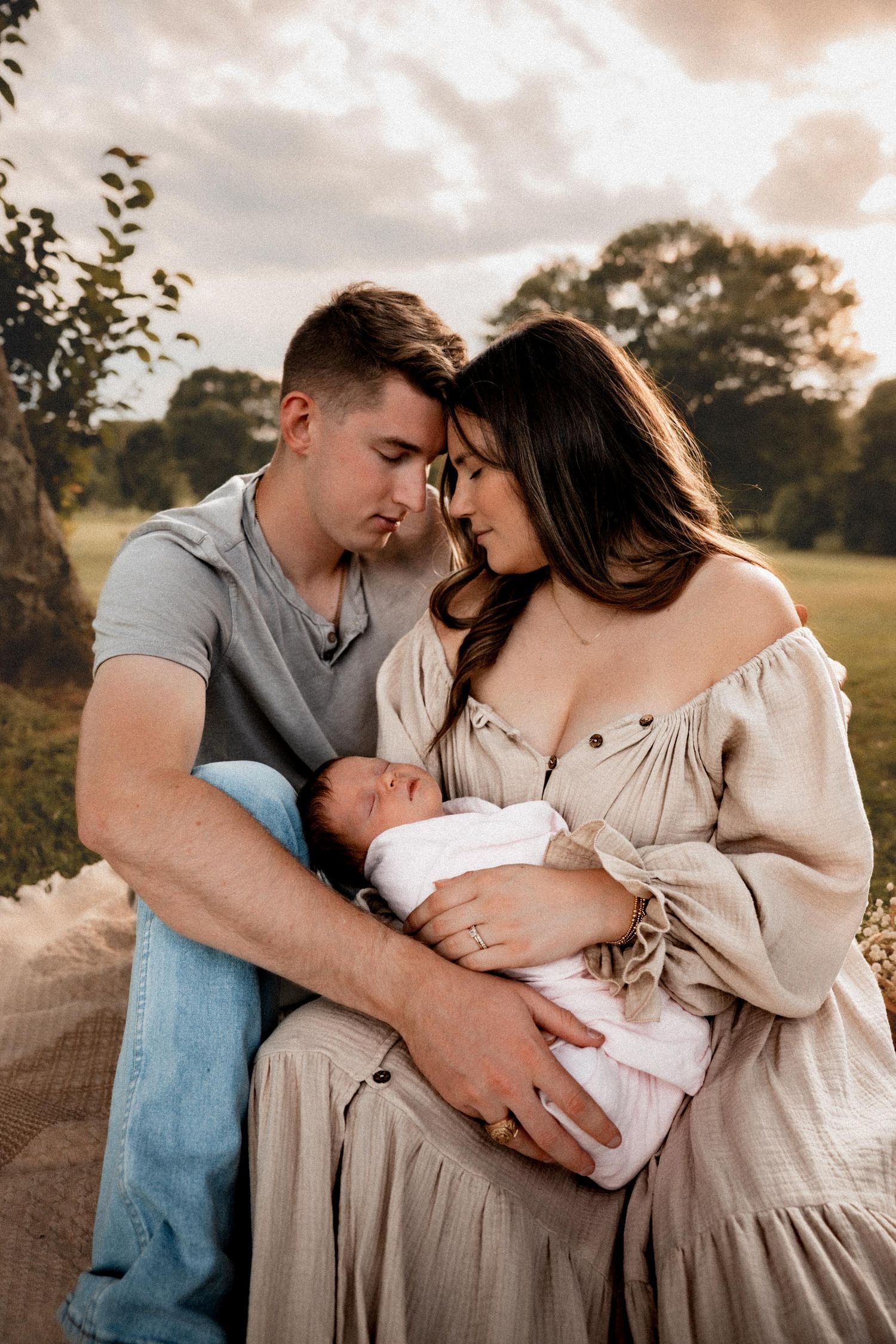 A couple tenderly cradles their newborn baby outdoors at golden hour, touching foreheads in an intimate family portrait.