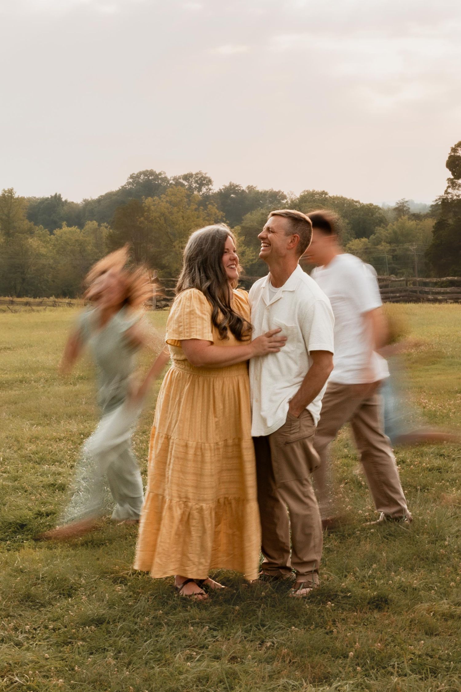 A couple embraces and laughs in a sunny field while blurred figures move around them during an outdoor photo session.