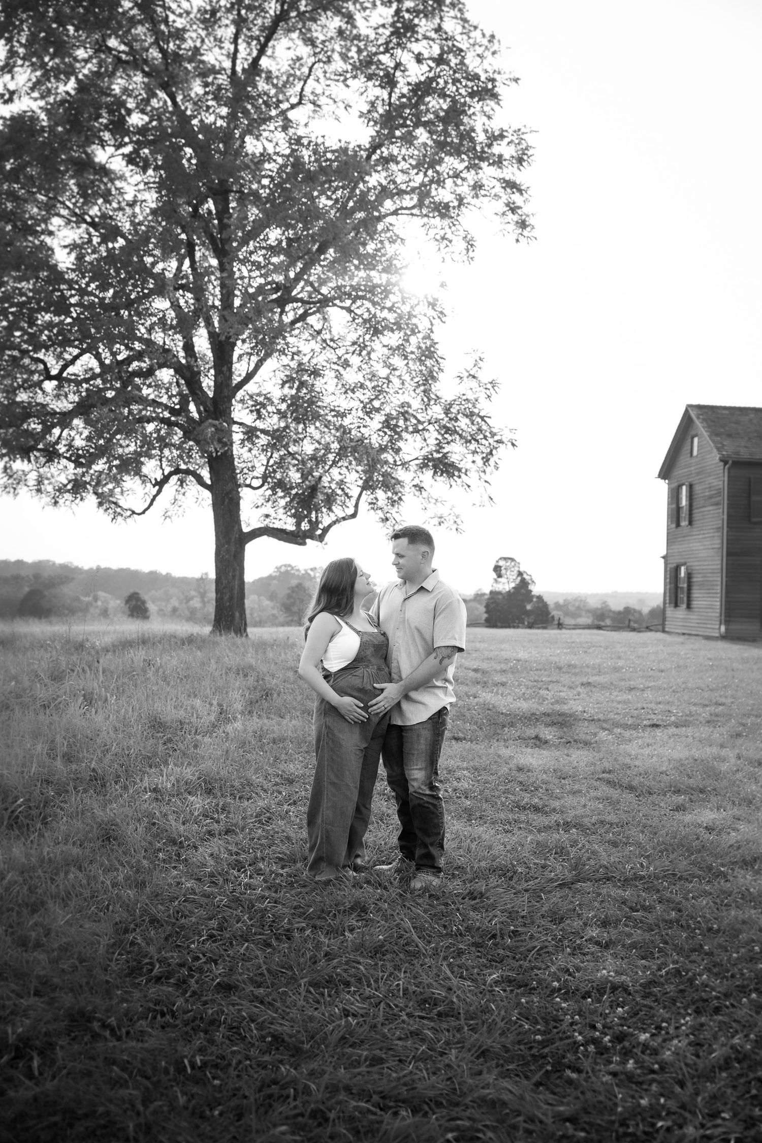 Black and white photo of a couple embracing in a field near a large tree and farmhouse at dusk.