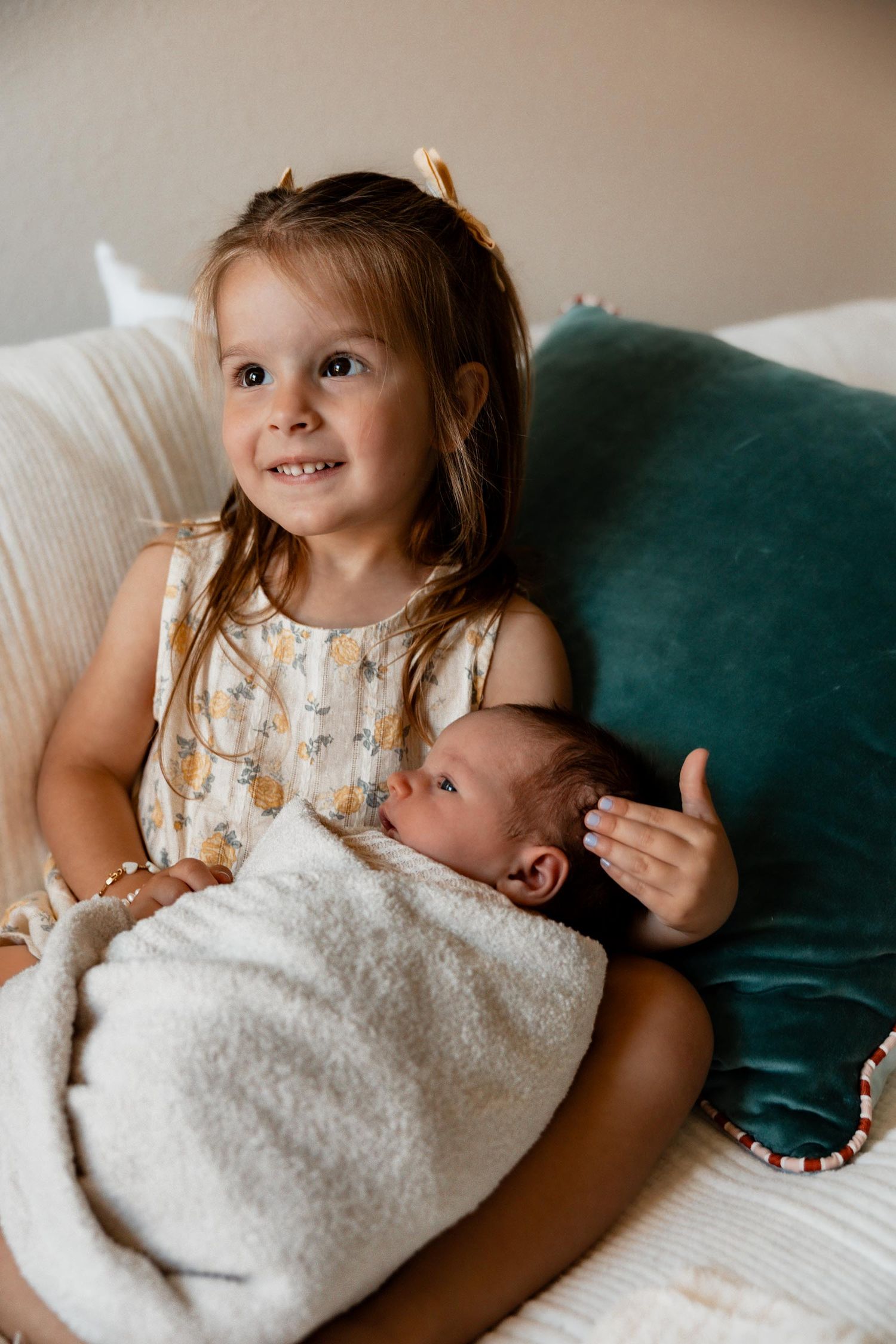 Smiling young girl with hair bows proudly holding swaddled newborn baby on a white couch.