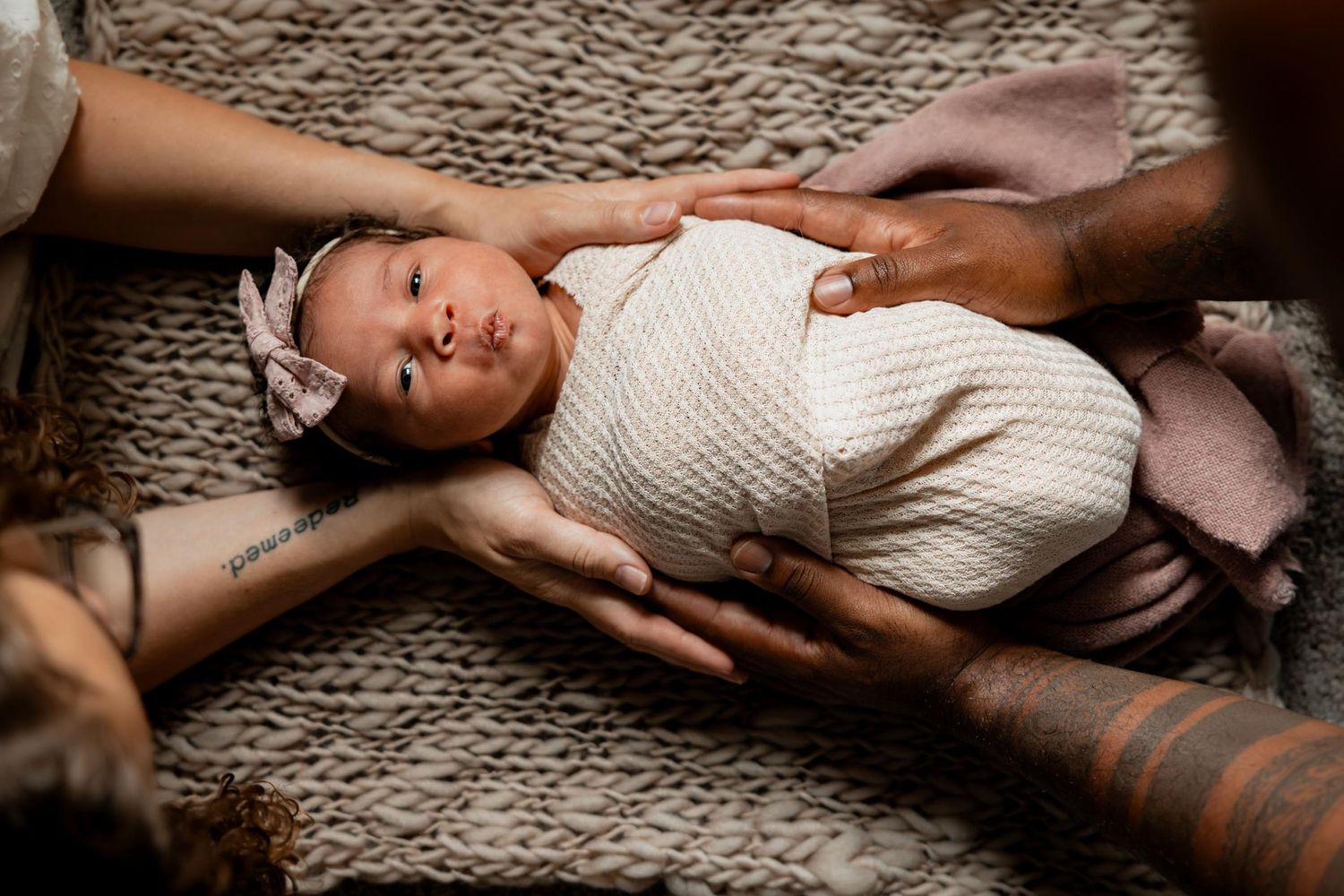 Newborn baby wrapped in cream knit swaddle with pink bow headband, cradled by two adults hands on a woven blanket.