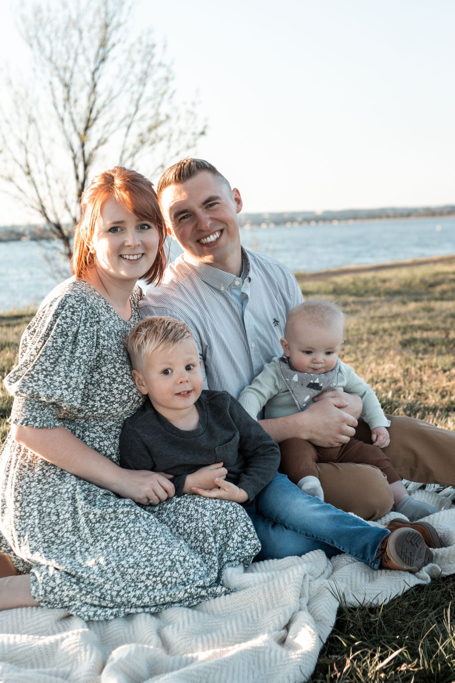 A family sits together on a blanket by a lake during a sunny outdoor photoshoot.