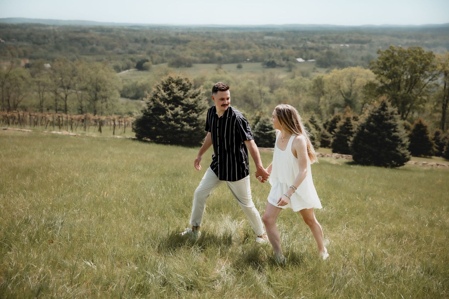 A couple holds hands and walks through a lush green field with trees and rolling hills in the background.