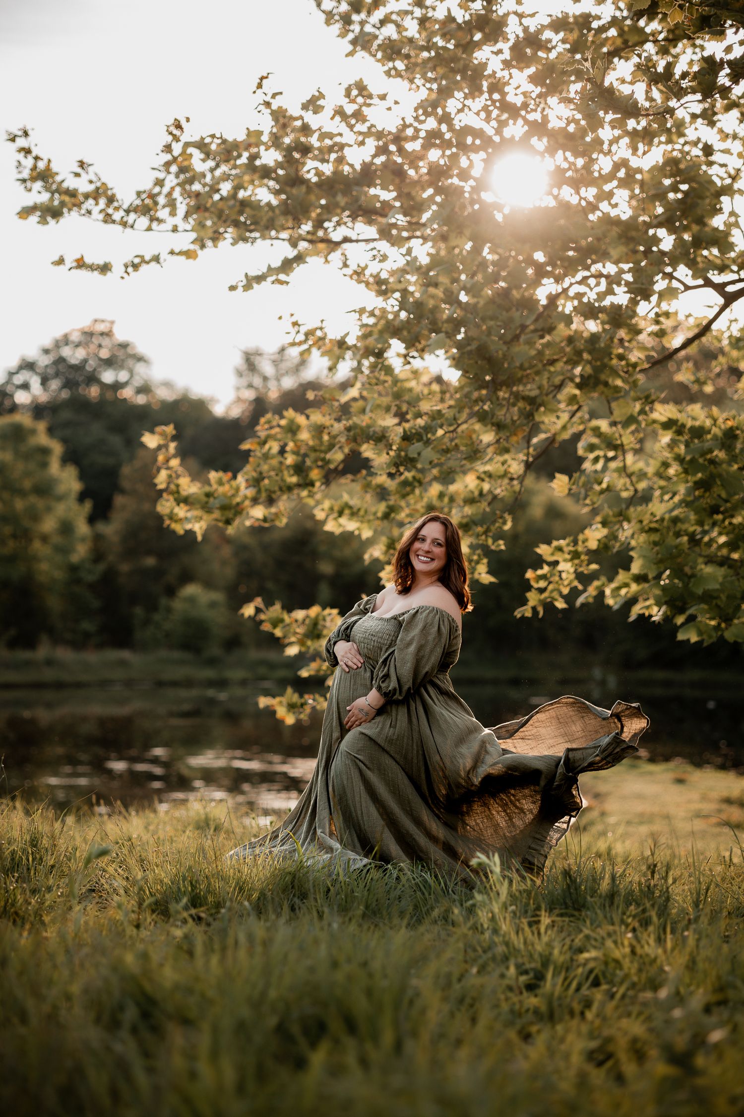 Pregnant woman in flowing olive green dress poses in sunlit park with golden hour light filtering through trees.