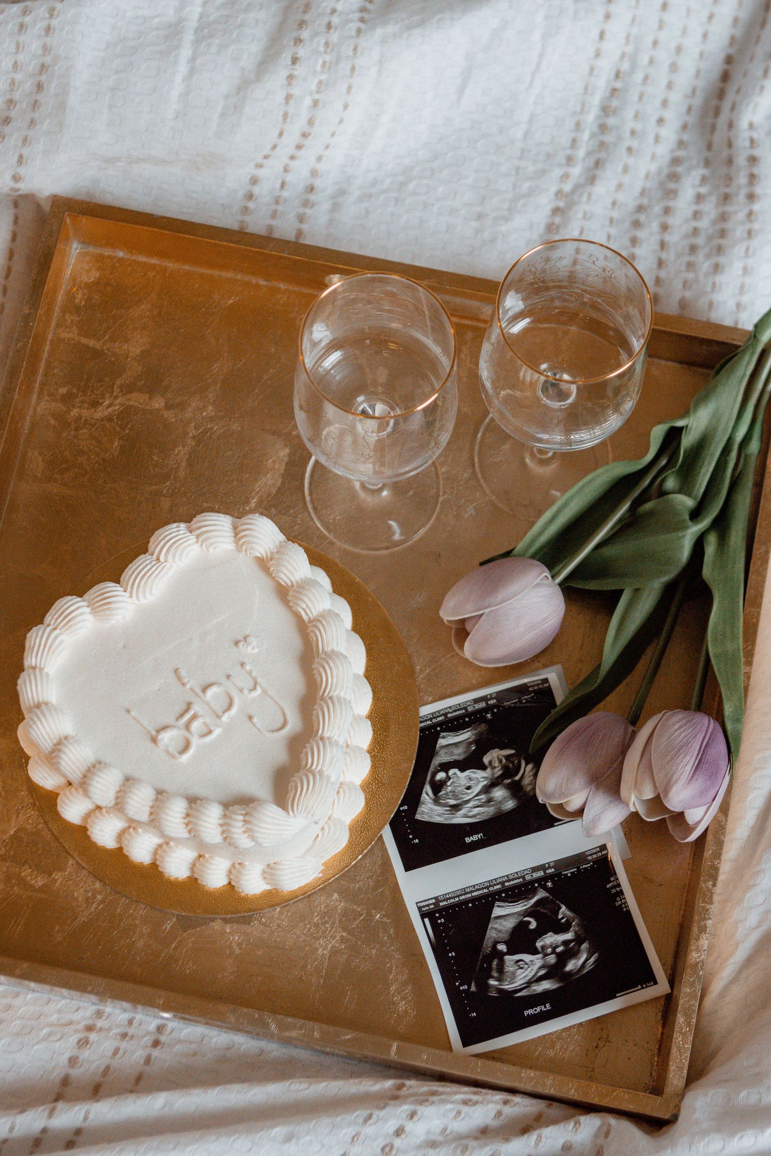 Heart-shaped white cake displayed alongside sonogram photos, wine glasses, and pink tulips on a wooden surface.