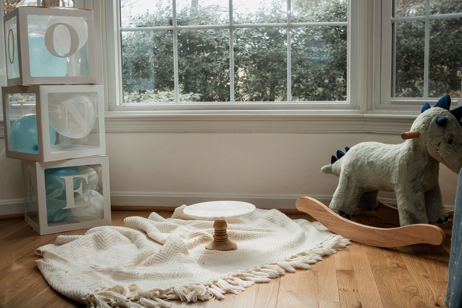 Cozy nursery corner with rocking horse, storage cubes, and white blanket on hardwood floor by large window.