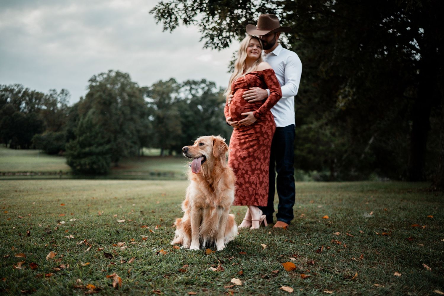 A couple and their golden retriever pose together in a grassy field during sunset.