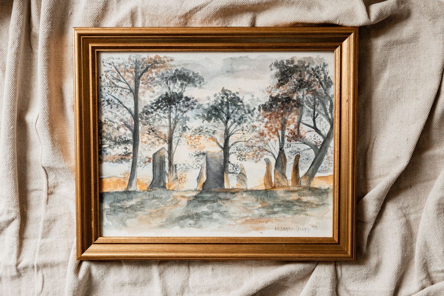 Three framed watercolor landscape paintings displaying forest scenes with trees hang on a cream-colored textile wall.