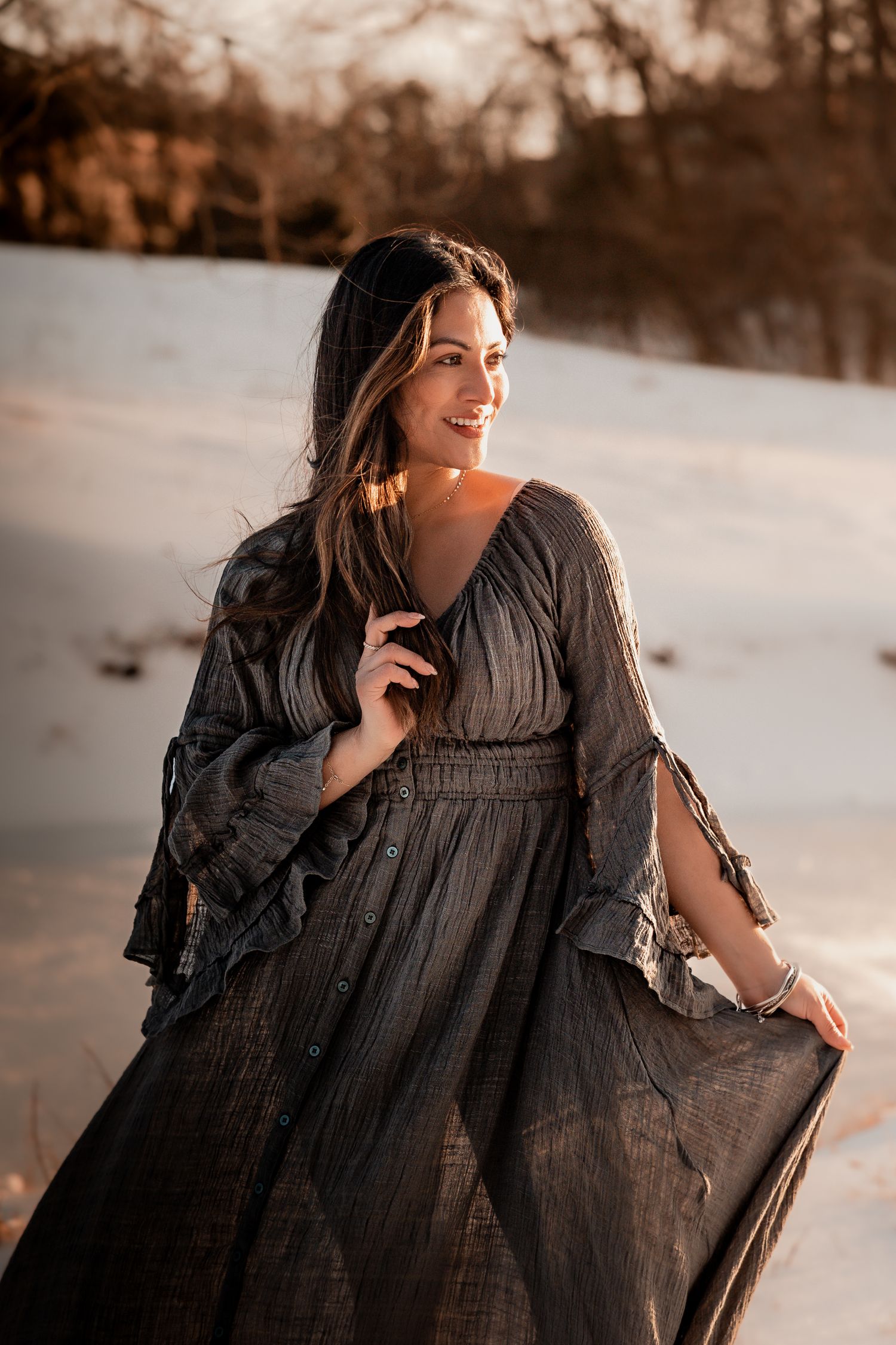 Woman in flowing dark dress poses outdoors in golden sunlight against snowy winter landscape.