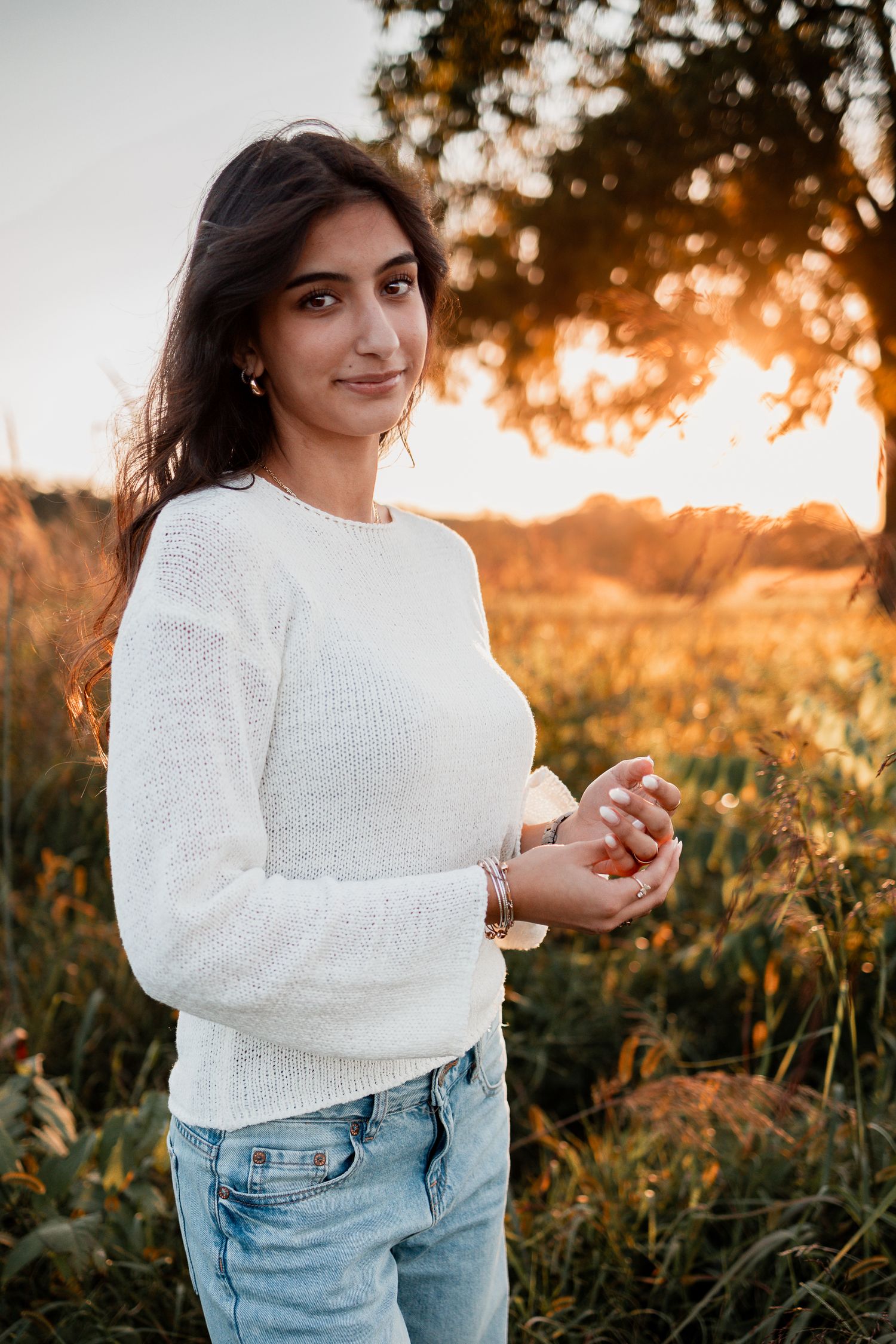 Woman in white sweater and jeans poses during golden hour sunset in tall grass.