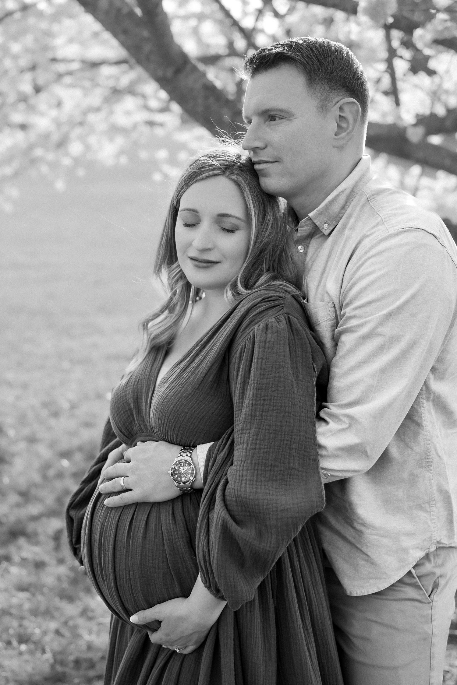 Black and white maternity photo of a couple embracing outdoors under a blossoming tree, man holding pregnant woman's belly.