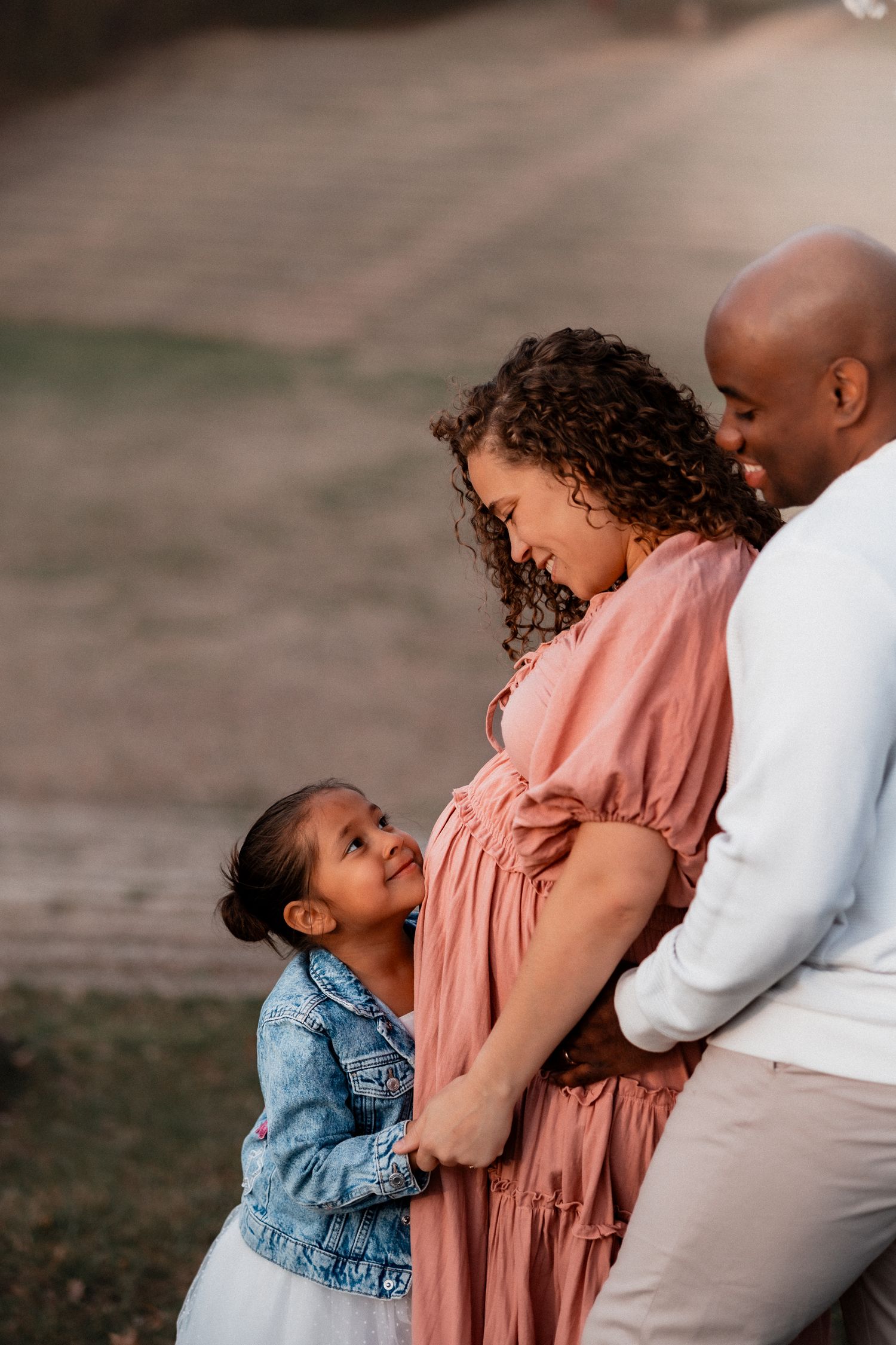 Pregnant woman in pink dress with curly hair embraces young daughter and bald husband outdoors during golden hour.