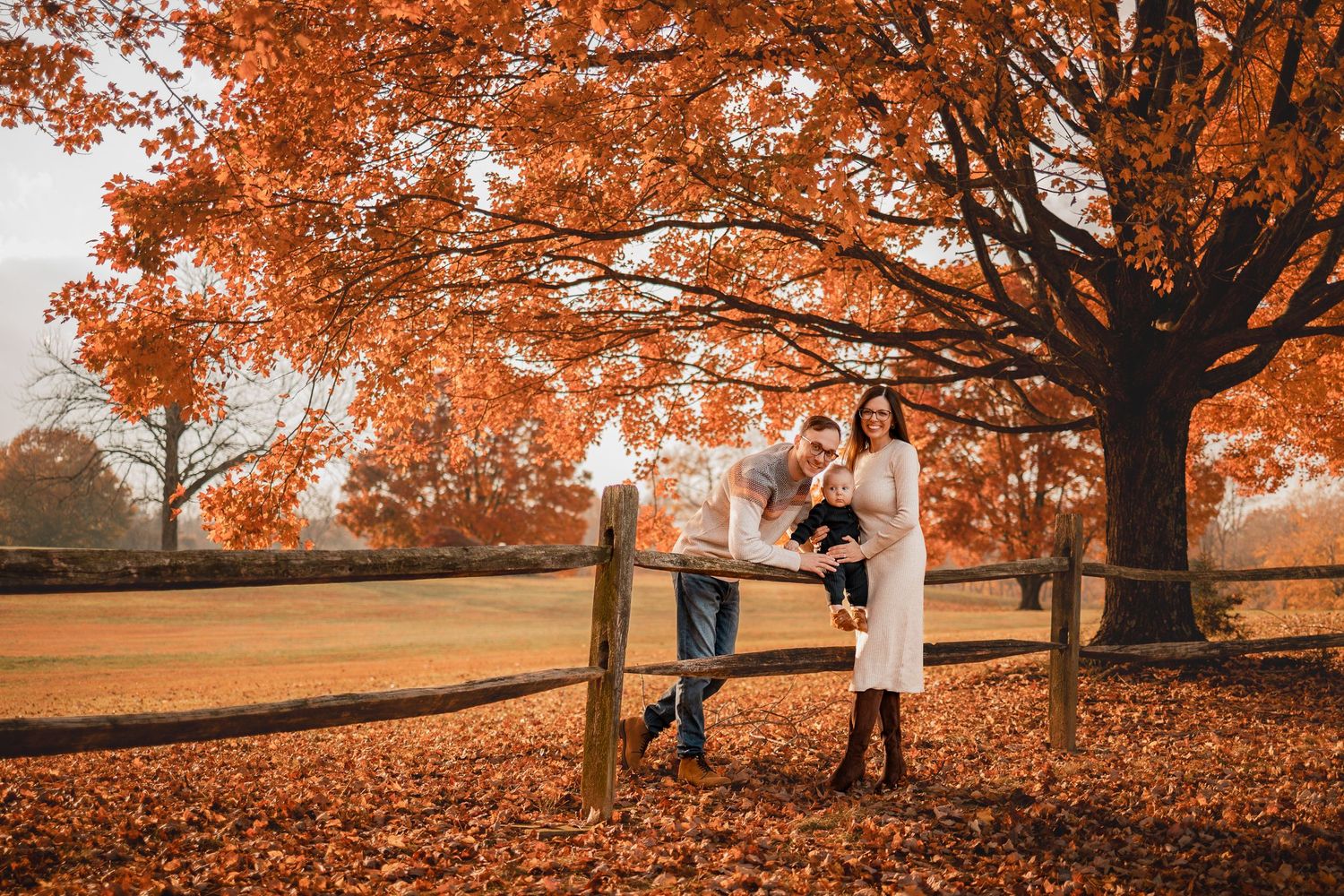 Family posing by wooden fence under vibrant orange autumn maple trees covered in fallen leaves.