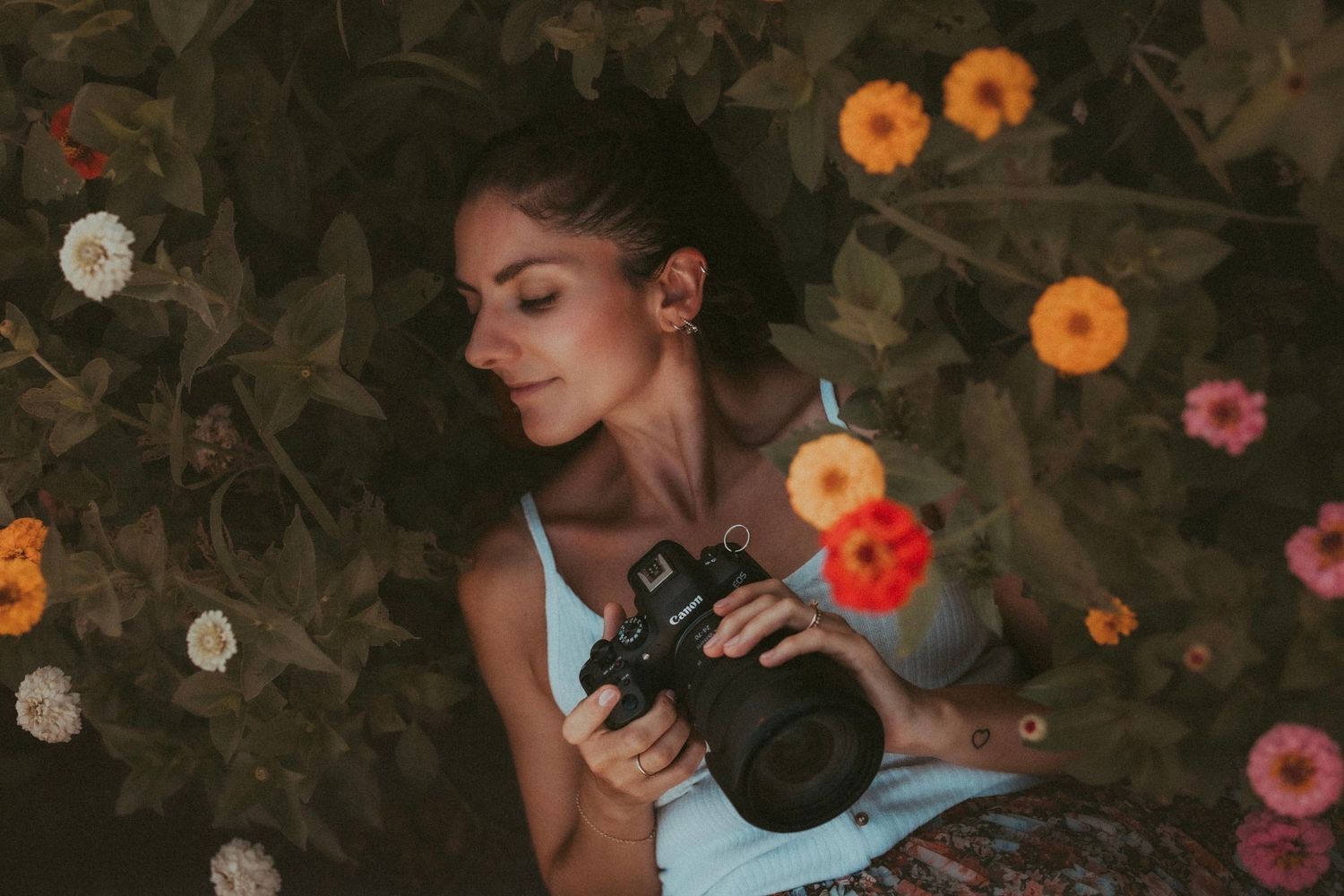 Woman lying among colorful flowers holding a camera, surrounded by lush green foliage in a serene outdoor setting.