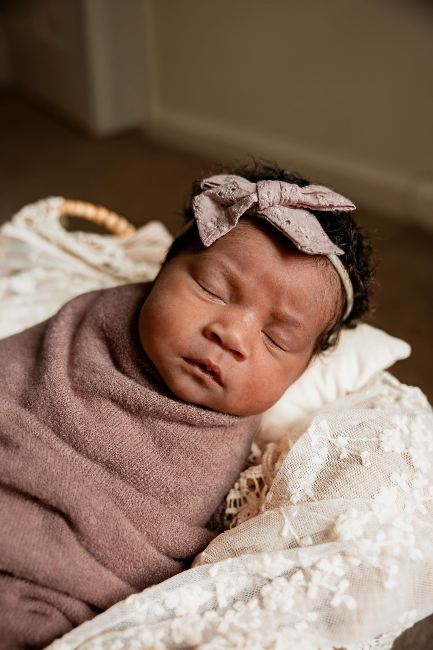 Sleeping newborn baby girl swaddled in mauve wrap with matching bow headband resting in a wicker basket.