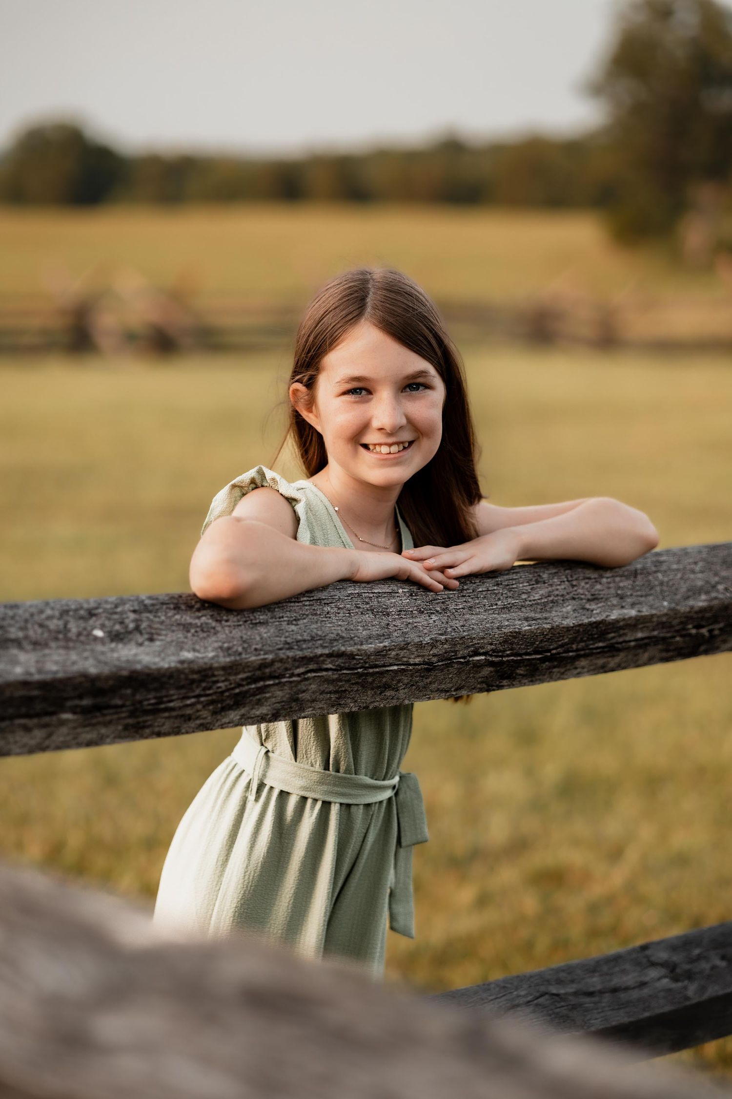 Smiling girl in mint dress leaning on rustic wooden fence with green field in background.