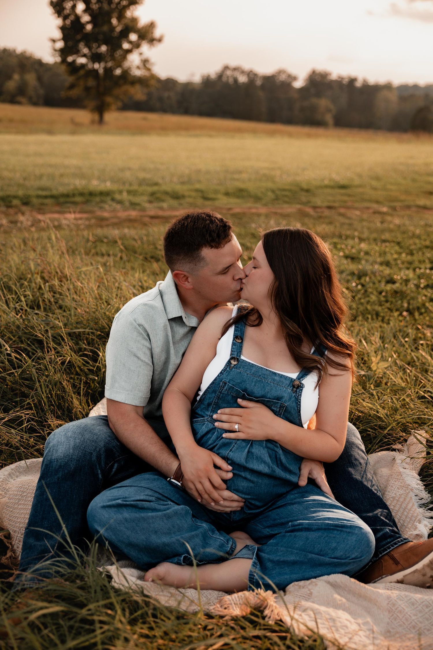 Couple sharing a kiss during golden hour maternity photo shoot in a sunlit field, woman wearing denim overalls.