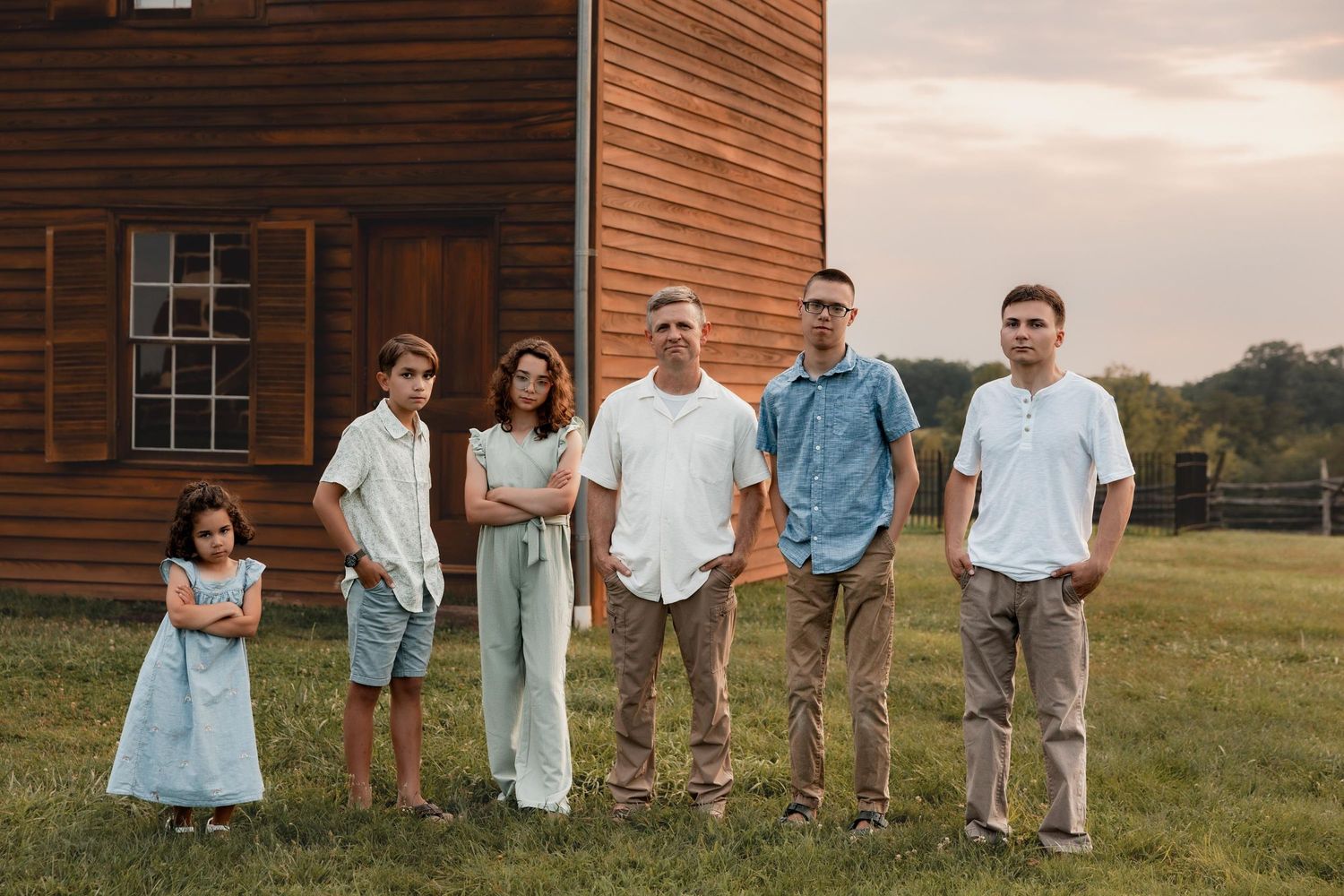 Family of six posing outdoors near a rustic wooden barn on a green lawn at dusk.