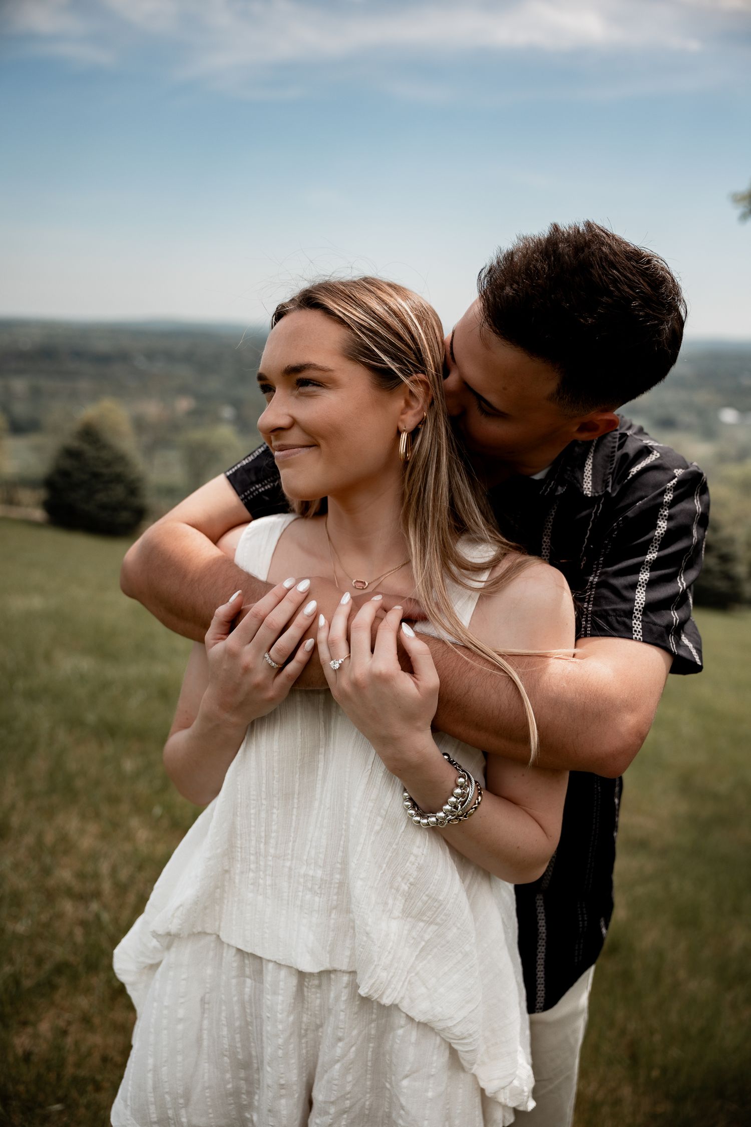 A couple shares a tender moment outdoors, man kissing woman's cheek while embracing her in a lush green landscape.
