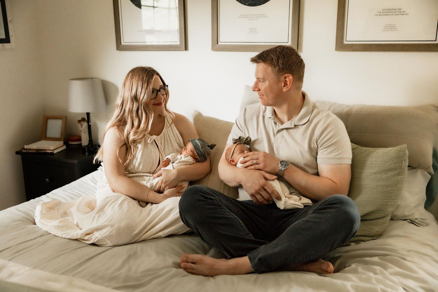 A couple sits on a bed holding newborn babies, smiling at each other in a cozy, warmly decorated bedroom.