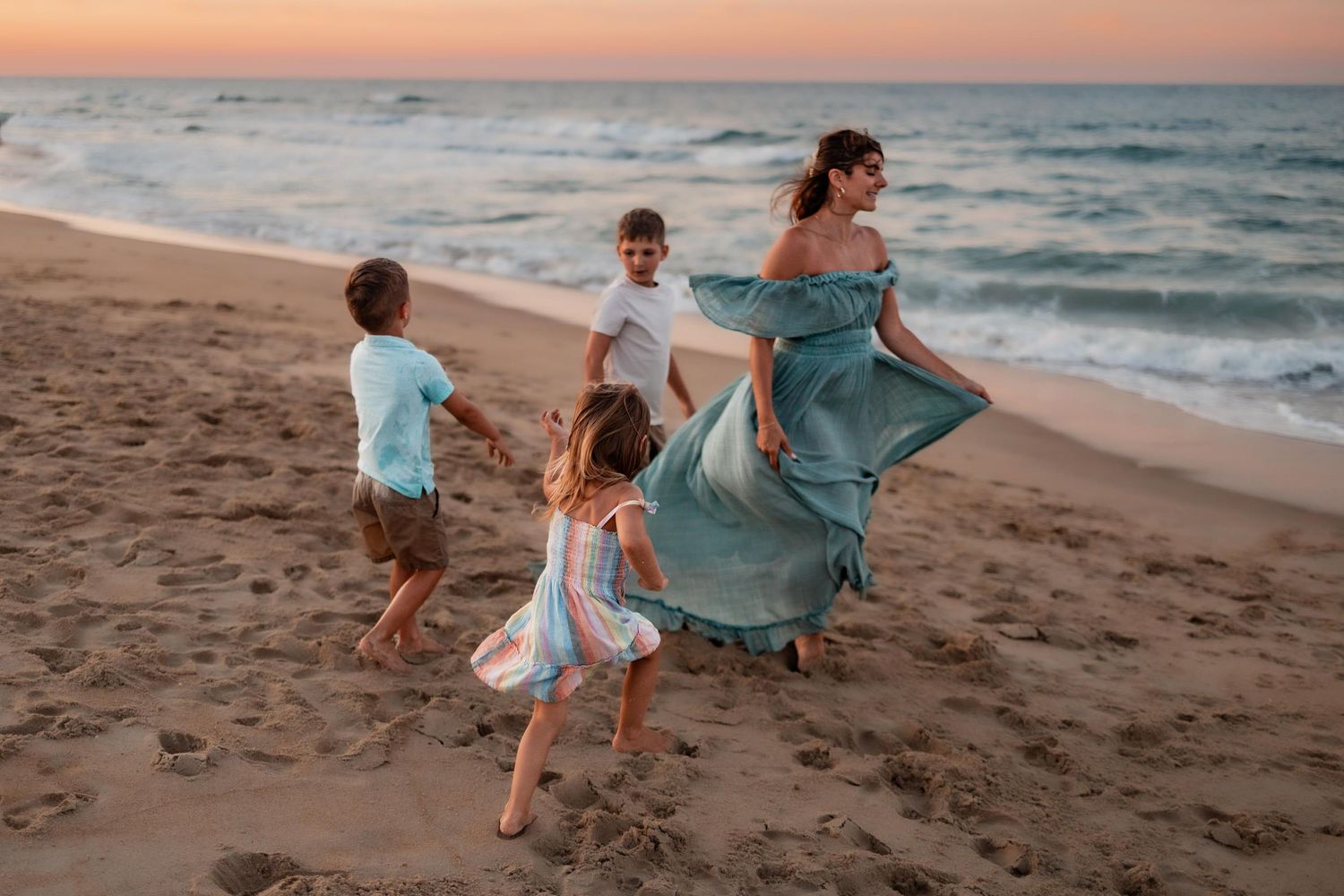 A mother in a teal dress dances with her three children on a sandy beach at sunset.