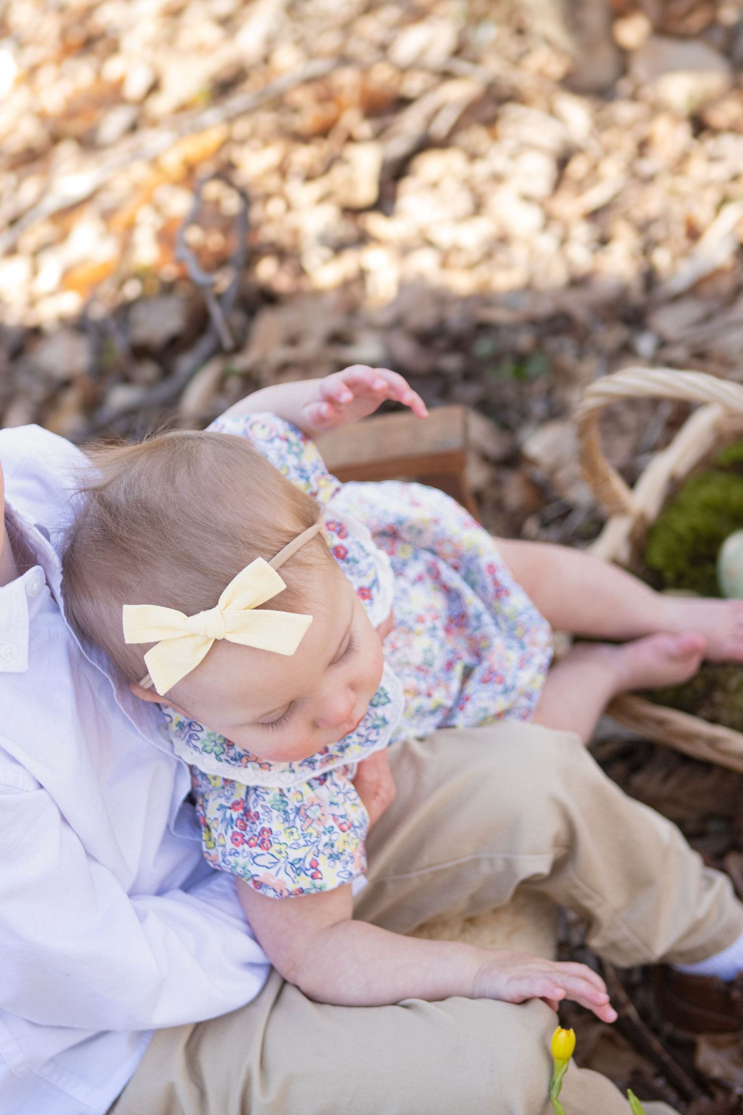 Baby in floral dress with bow examines surroundings while sitting outdoors.