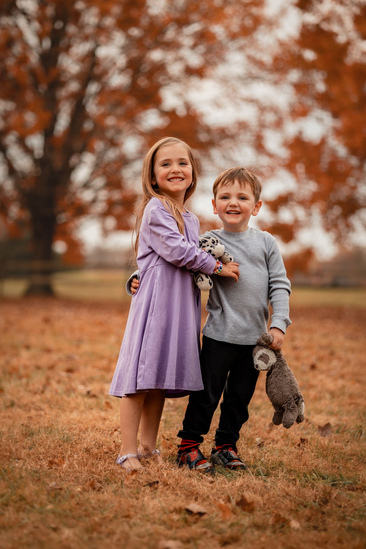 Two children in purple dress and grey sweater hold stuffed animals while standing on fallen autumn leaves.