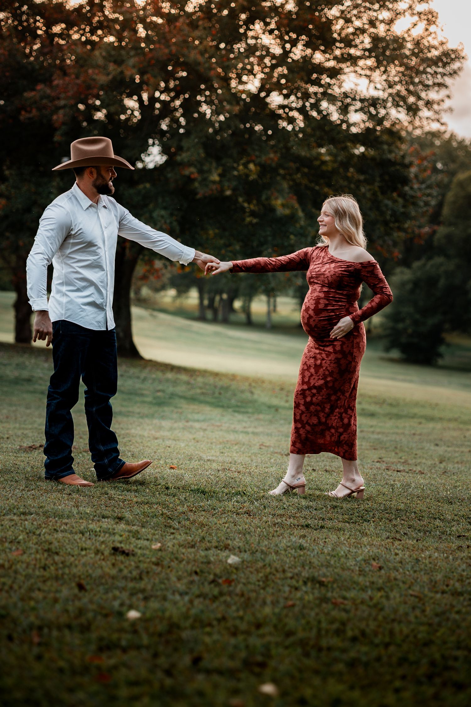 A couple dances playfully on a grassy hillside during sunset, with one person wearing a flowing red dress.