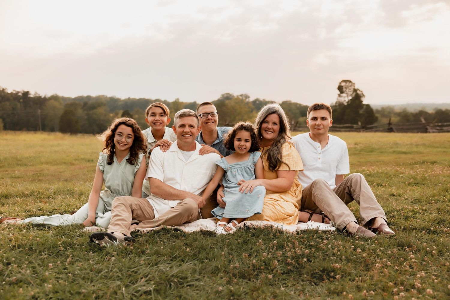 A joyful family of eight poses together on a blanket in a lush green field during golden hour.