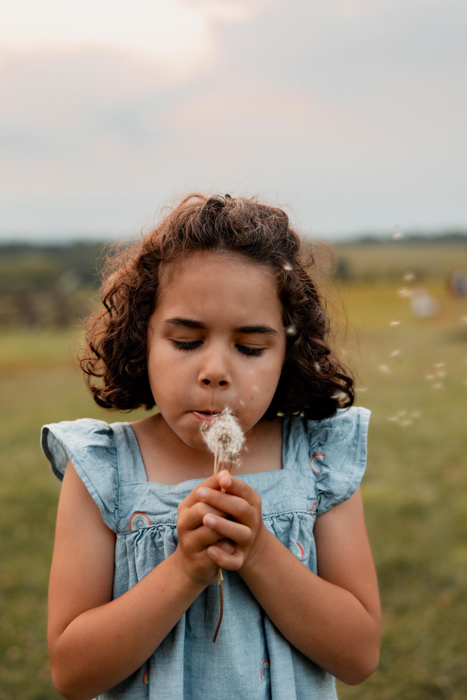 A young girl with curly hair blowing dandelion seeds in a green field, wearing a blue ruffled dress.