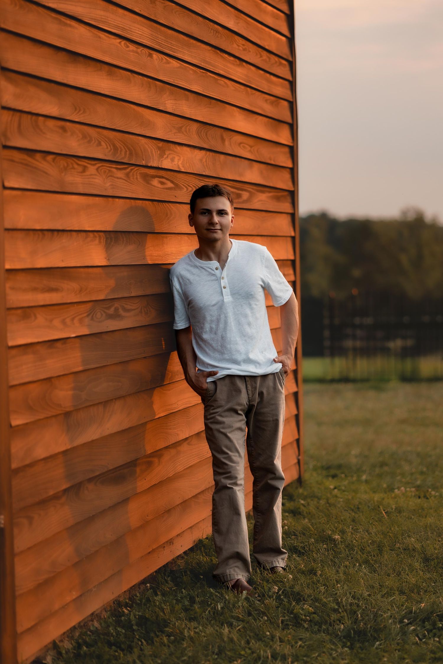 A young man in a white shirt and khaki pants leans against a warm wooden wall at sunset.