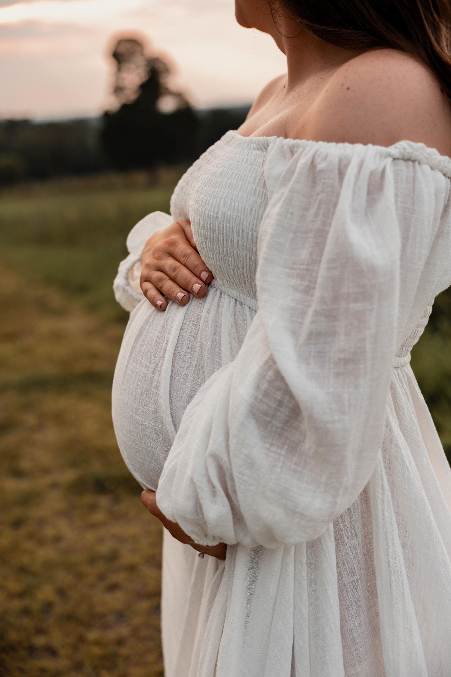 Close-up of pregnant woman in white off-shoulder dress cradling her baby bump at sunset in a green field.