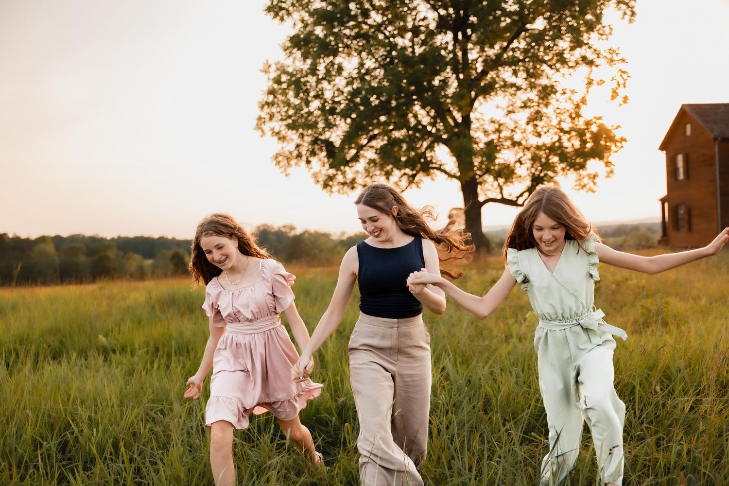 Three girls hold hands and walk through a lush green field at sunset near a large tree.